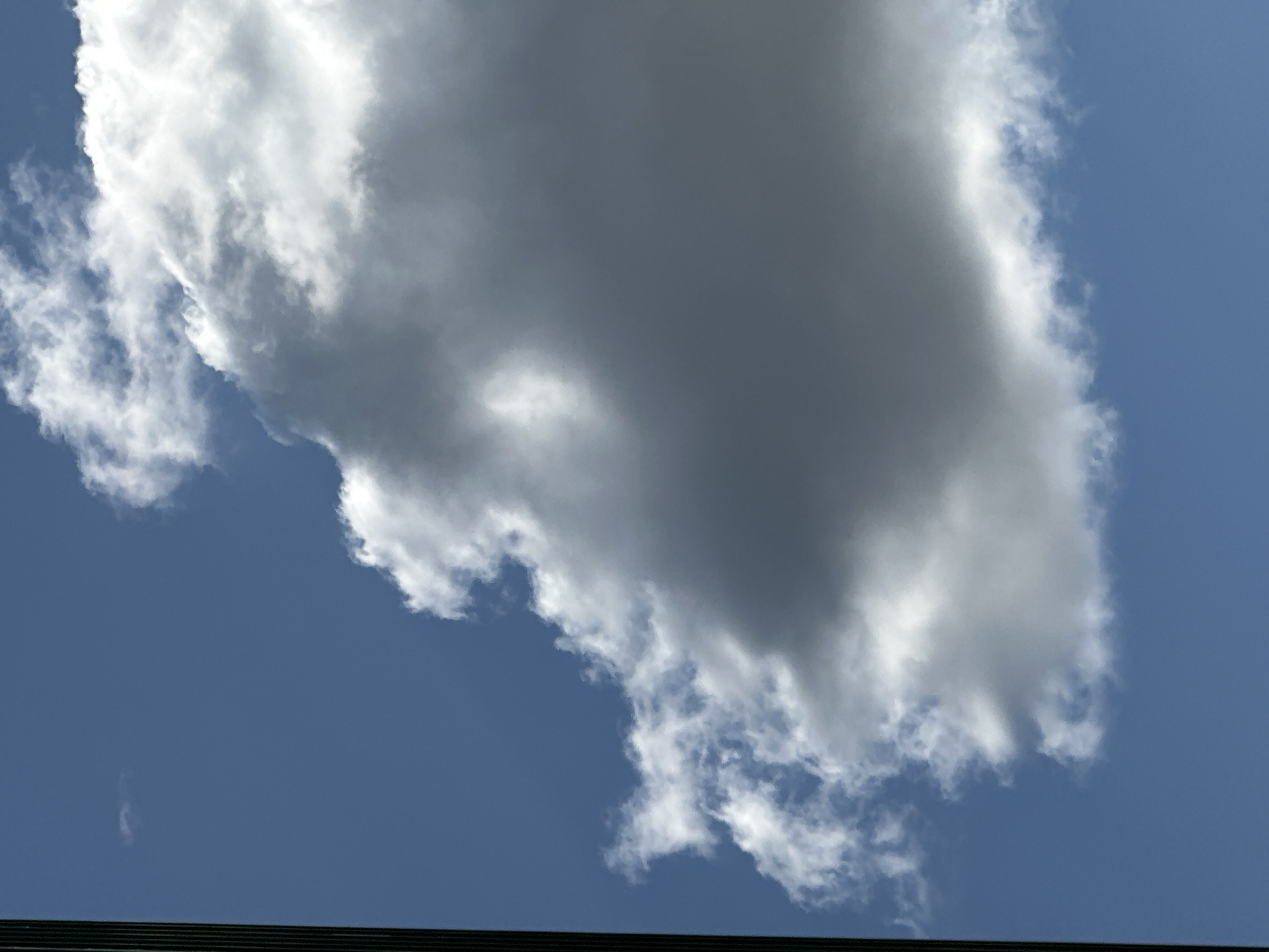A full-frame, portrait-oriented photograph captures a vast expanse of blue sky. A large, dark gray cloud dominates the right side of the frame, its top portion transitioning to bright white where it catches the sunlight. To the left of this prominent cloud, a scattering of smaller, wispy white clouds drifts against the blue backdrop. The left edge of the image features a thin, vertical dark gray bar, likely the edge of a structure or an element of the photo frame. The overall scene is serene and natural, with no discernible signs of human activity or urban elements.
