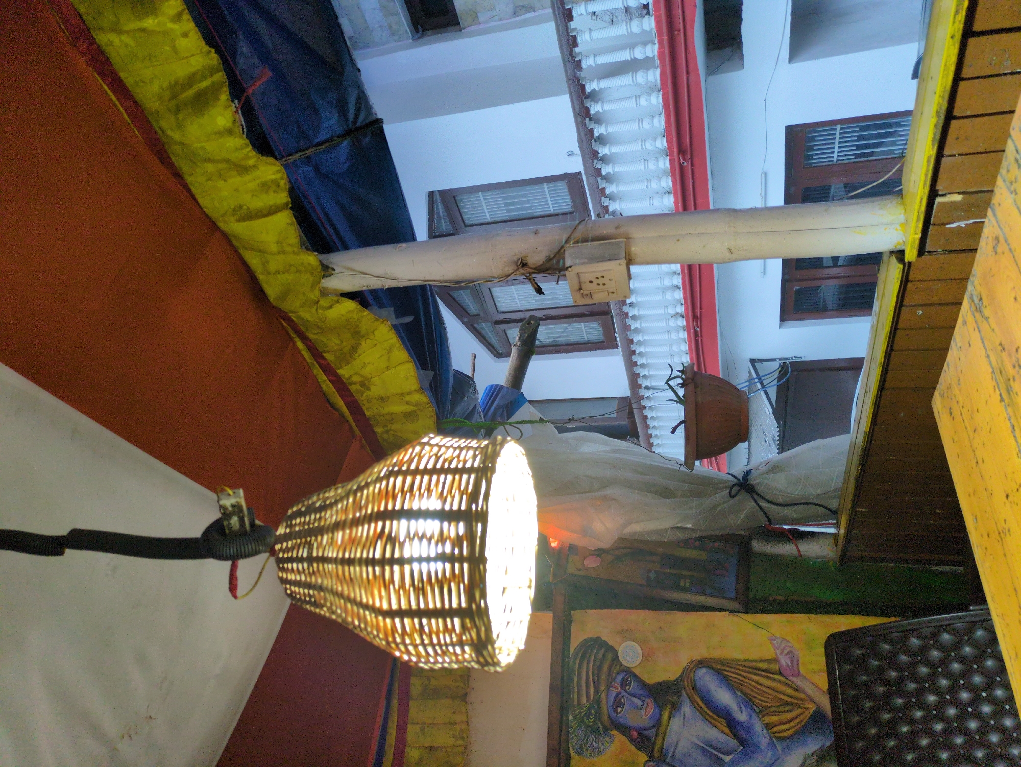 The image depicts an outdoor covered seating area, likely at a restaurant or cafe, in Bhagsu, India. The focus is on a hanging woven lampshade emitting a warm light, suspended from a black electrical conduit attached to a white ceiling. To the left, a colorful painting of a figure with a blue face and elaborate headdress is visible, with a black tufted chair in the foreground. Beyond this, a white building with a red-trimmed balcony and multiple windows with iron grills is seen. A brown terracotta pot hangs from a pole to the right of the painting, and a section of a yellow wooden table is in the foreground. The overall impression is one of a rustic, possibly bohemian, setting.