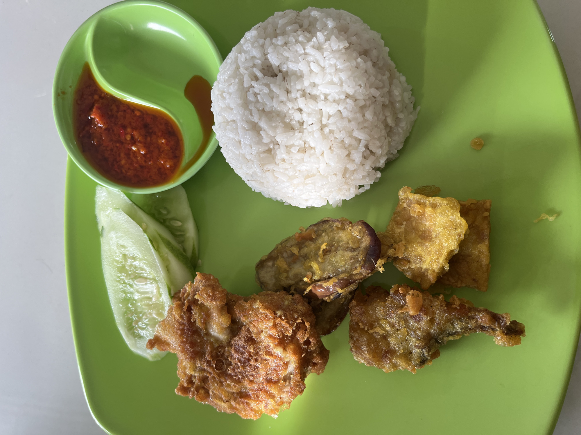 A plate of Indonesian food, possibly nasi uduk or nasi campur, is presented on a bright green plate. The meal consists of a mound of white rice, several pieces of golden-brown fried chicken or tempeh, slices of cucumber, and a small dish of red chili sauce. The food is arranged on a white surface, which could be a table. The image is taken from a top-down perspective. There is no indication of the city of Bireuenmeunasahreuleut, Indonesia, or any people in the image.