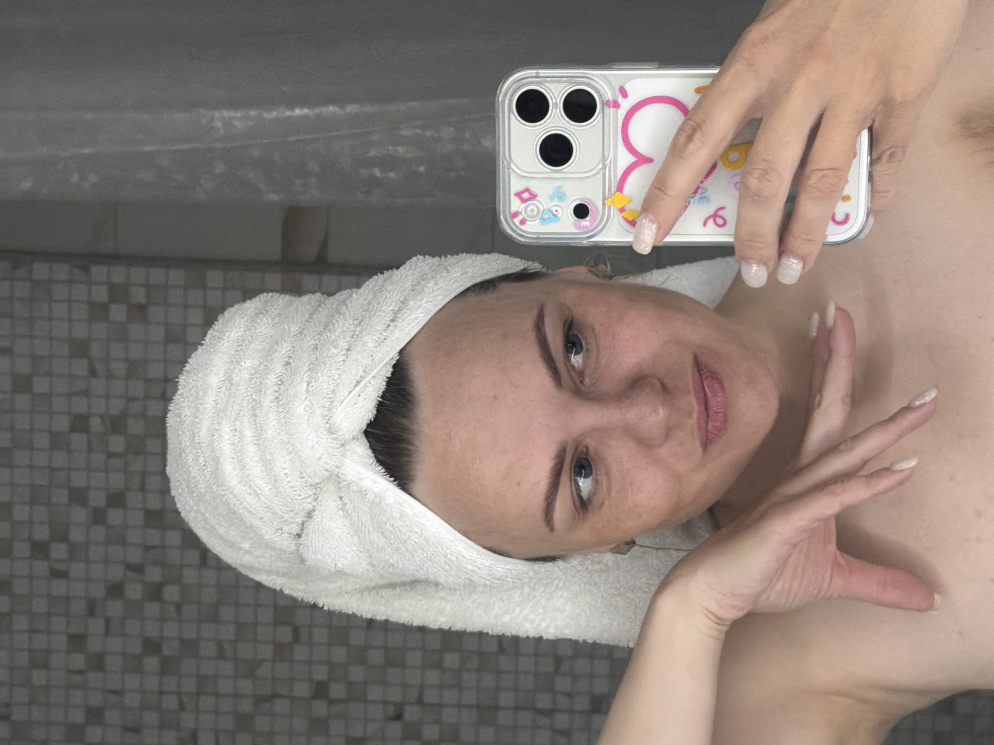A person with fair skin and a white towel wrapped around their head is taking a selfie in what appears to be a bathroom. The person is holding a smartphone with a decorative case in their right hand. The bathroom has gray and white tiled walls. There is no information available to verify or deny that the location is in Votkinsk, Russia.