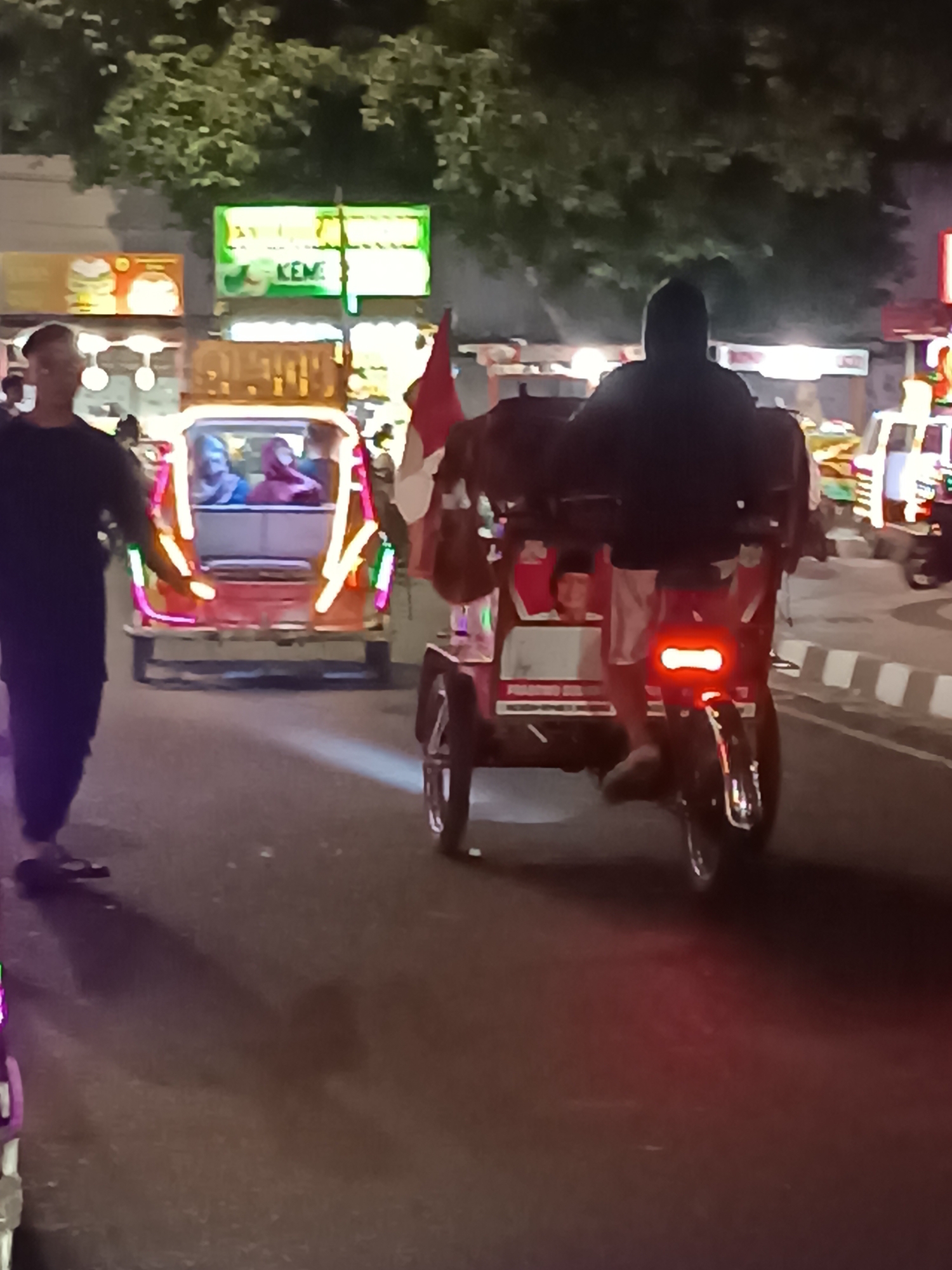 The image depicts a street scene at night in Yogyakarta, Indonesia, likely a commercial or entertainment district. In the foreground and center, a person wearing a dark hooded jacket is riding a brightly lit three-wheeled vehicle, possibly a modified bicycle or motorcycle, commonly known as an "odong-odong" or "becak motor." The vehicle is decorated with colorful LED lights and has a red rear brake light illuminated. To the left, another person in dark clothing is walking on the street, facing away from the camera. Behind the person on the vehicle, a passenger vehicle, likely an "odong-odong" as well, is visible with passengers inside and is also decorated with lights. In the background, neon signs are illuminated, suggesting the presence of shops or food stalls. The street appears to be paved, with a raised sidewalk visible on the right side. Trees with dense foliage are present above the buildings and signs. The overall atmosphere suggests a lively evening setting.