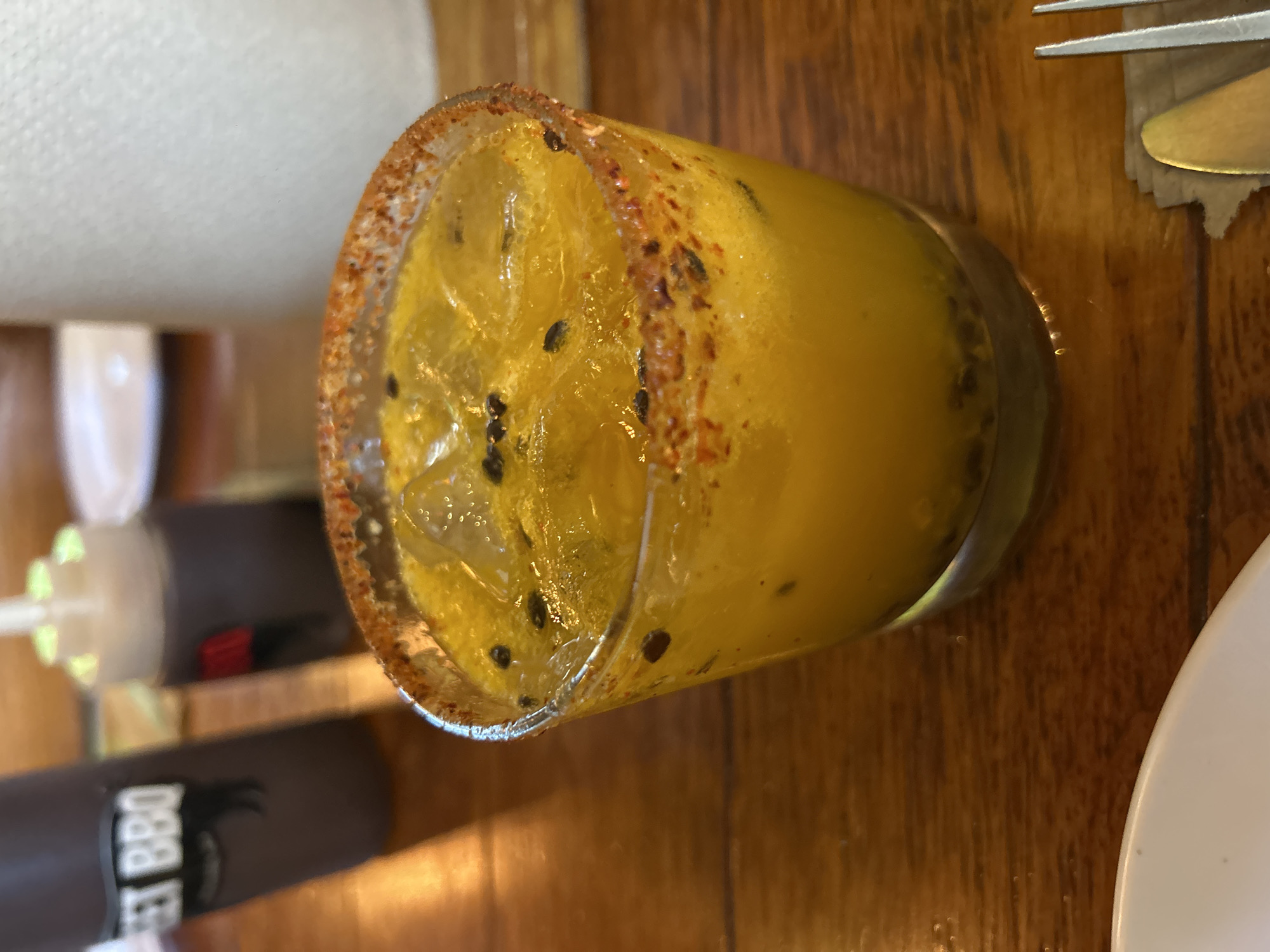 The image shows a partially consumed cocktail on a wooden table. The cocktail is in a clear glass with a rim coated in a reddish-brown seasoning. The drink itself is yellow and contains ice and what appear to be small black seeds. Next to the glass, there are cutlery items and a white plate. In the background, there is a bottle of Sweet BBQ sauce. The location is not discernible from the image.