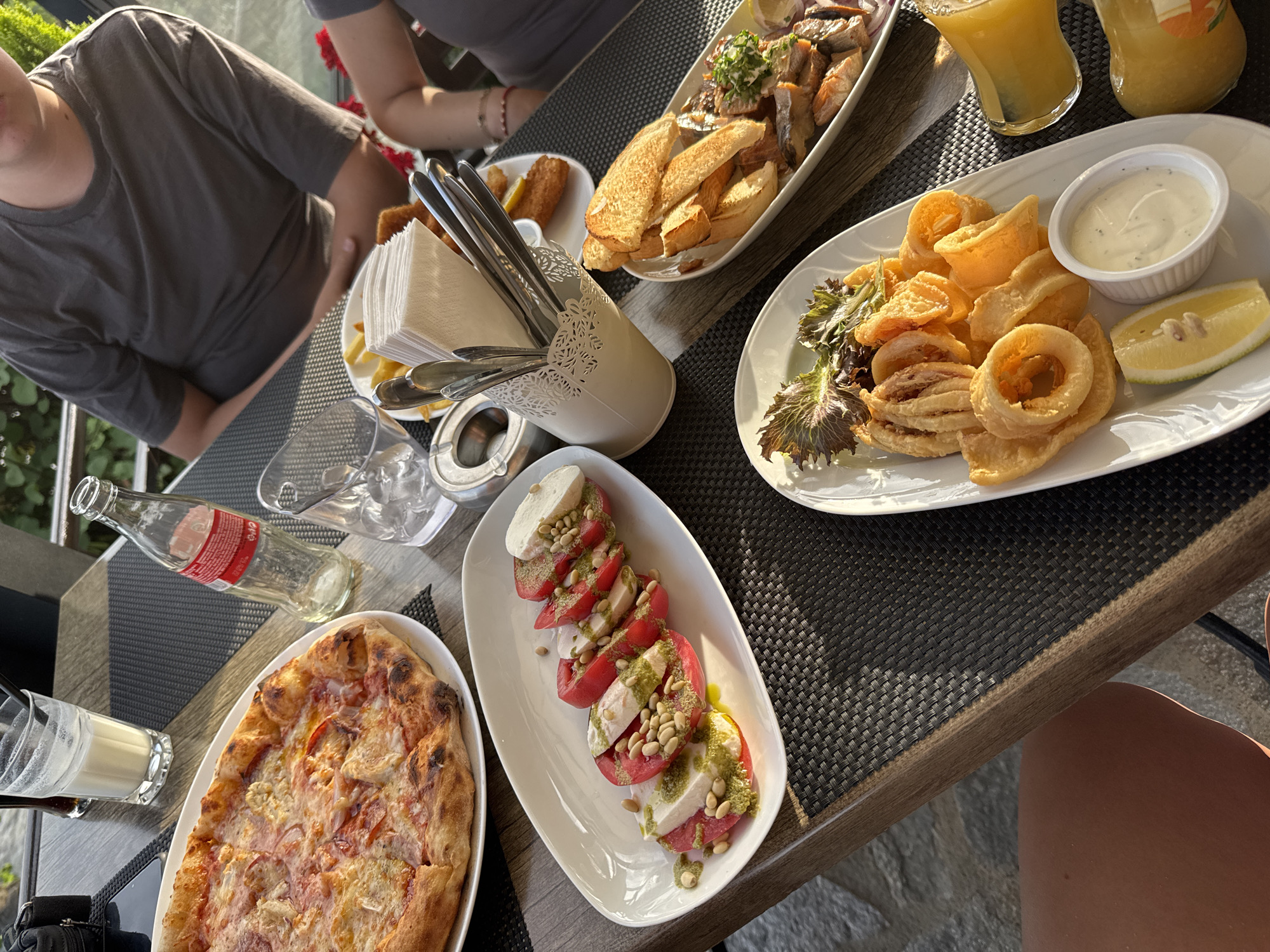 An outdoor dining scene unfolds on a table with a textured dark placemat. A pizza with melted cheese and red sauce is a prominent dish. Adjacent to it, a white plate holds a Caprese salad with sliced tomatoes, fresh mozzarella, pine nuts, and a green sauce. Another plate features fried calamari rings with a side of white dipping sauce and a lemon wedge. In the background, a plate of fish and bread, fries, and a glass of orange juice are visible. A person in a grey t-shirt is seated at the table. The setting appears to be a restaurant in Sozopol, Bulgaria, indicated by the presence of local currency (which is not visible in this specific image, but implied by context) and the overall ambiance suggestive of a seaside town. The lighting suggests it is daytime, likely late afternoon, with sunlight casting shadows.