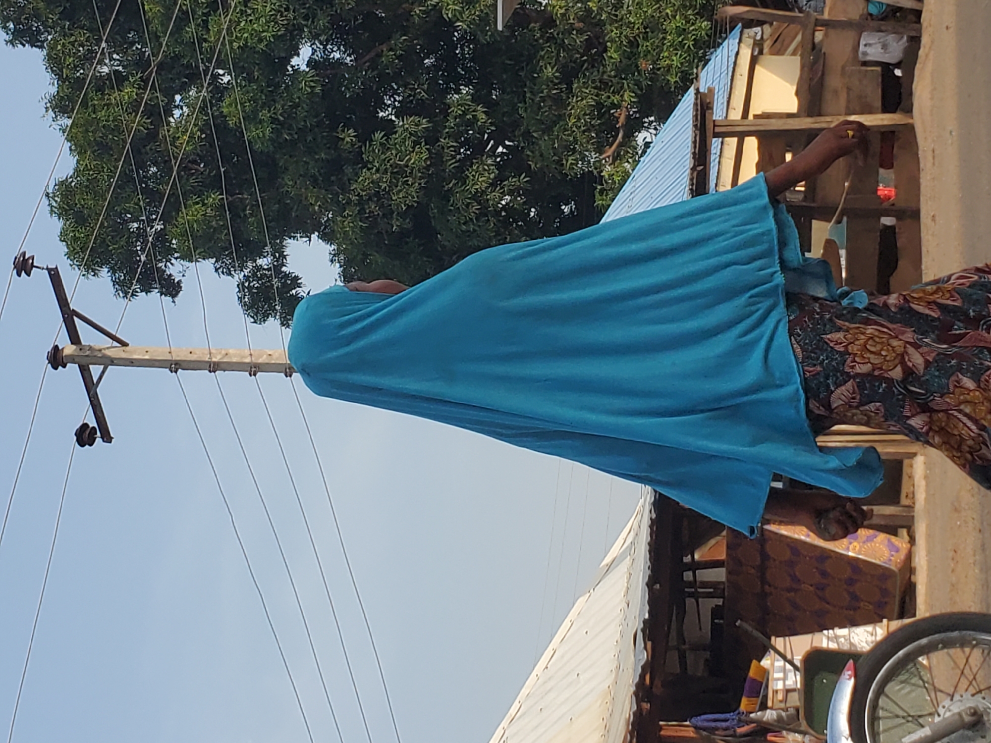 A woman wearing a blue garment is standing in Mararaba, Nigeria. She is near a utility pole and power lines. A building with a corrugated metal roof and wooden supports is in the background. A motorcycle is parked nearby, and various household items are visible in the cluttered area.