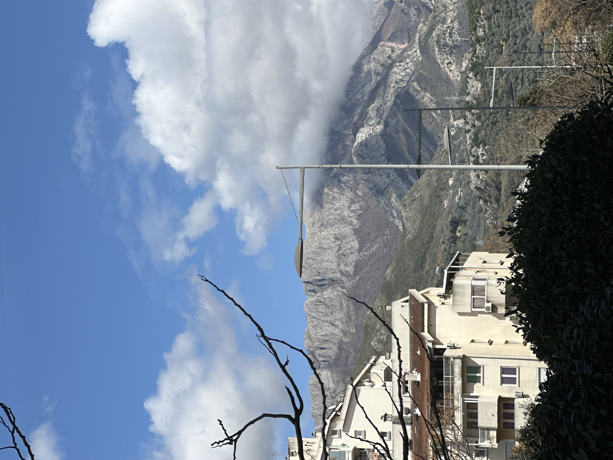 A daytime scene presents a building set against a backdrop of mountains and sky. The building, located in Šušanj, Montenegro, appears to be an apartment complex or multi-story residential structure. Parts of its facade are visible, featuring windows, balconies, and a light-colored exterior. A bush or tree with dark foliage is situated on the right side of the building. In the background, mountains rise steeply. Clouds partially obscure the peaks, contributing to an overcast feel. Utility poles are erected near the mountain and behind the building. In the foreground, bare branches of a tree extend into the sky, which is a mix of blue and clouds.