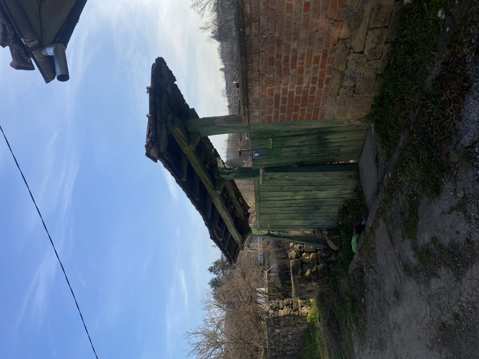 The scene depicts a rural residential entrance on a clear, sunny day in Dobri dial, Bulgaria. The primary subject is an weathered, dark green, two-panel wooden gate, slightly ajar, topped by an overhanging tiled roof supported by wooden beams. A small blue rectangular sign bearing the white numeral "9" is affixed to the right gate panel, with a smaller white rectangular object beneath it.
To the immediate right of the gate, a wall constructed with a red brick upper section rests upon a lower stone foundation or wall. Above this wall, a metal gutter spout is visible from a roof overhang. To the left of the gate, a dry-stacked stone wall, partially overgrown with vegetation, extends into the background.
The ground in the foreground consists of a dirt or gravel path with patches of dry grass and fallen leaves. A concrete slab forms a threshold at the gate entrance. A small green plastic bag is visible near the base of the left gate post. The background reveals bare deciduous trees and evergreen conifers, indicative of a dormant season, and glimpses of distant structures or houses. The sky is bright blue with minimal wispy clouds. A single dark utility wire traverses the upper left portion of the frame. No individuals or active interactions are present within the image.
