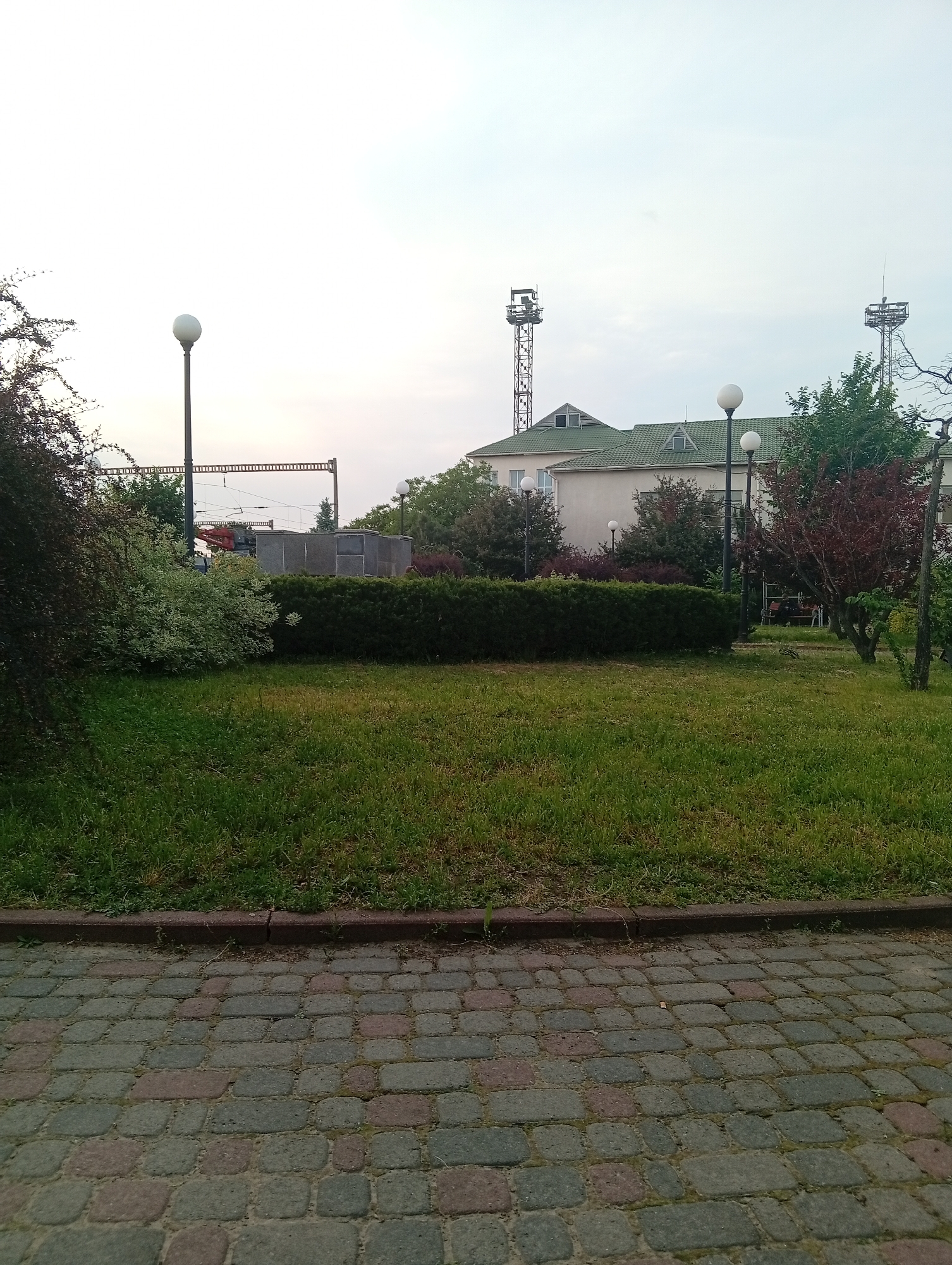 The scene shows a park-like area in Poltava, Ukraine, with a paved brick walkway in the foreground and a grassy area beyond. A long, neatly trimmed hedge runs horizontally across the midground, with various trees and bushes behind it. A building with a green roof and two communication towers on its roof is visible in the background. Three white globe streetlights are visible: one to the left and two to the right. Overhead electrical infrastructure, with a crane, is partially obscured by the trees on the left. The sky is overcast. No people are readily visible.