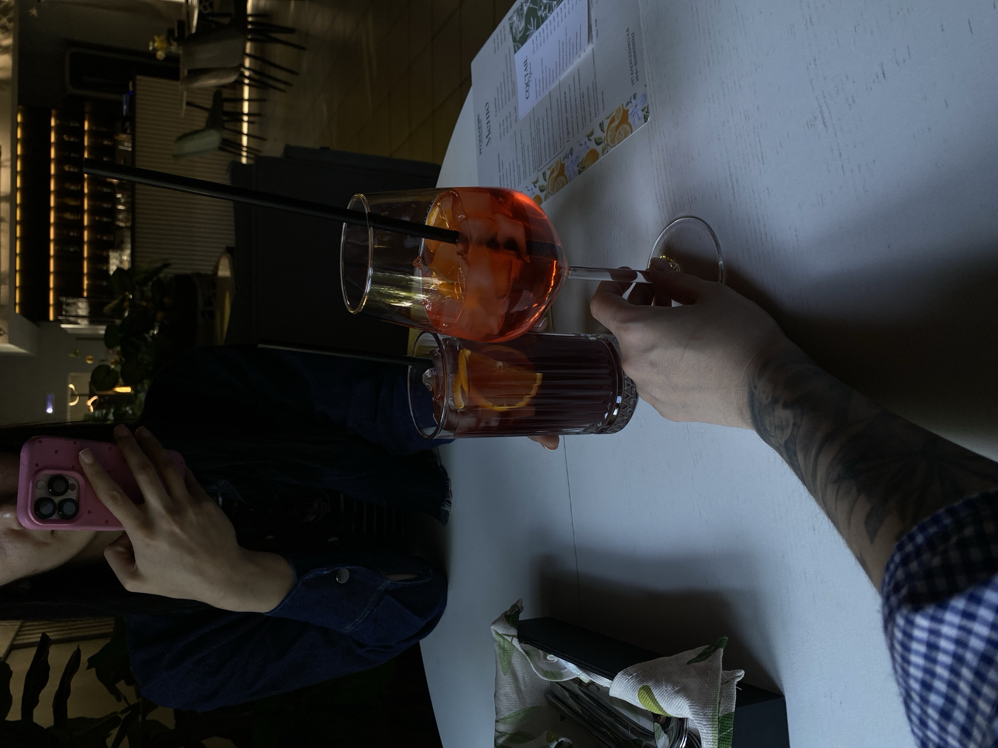 The image depicts an indoor scene, presumably in a restaurant or bar in Votkinsk, Russia. Two individuals are present, one taking a photo with a pink phone. The other person is holding two drinks: an orange cocktail in a wine glass, complete with ice and a straw, and another cocktail in a highball glass with a slice of orange inside. A menu is visible on the white tabletop along with some cutlery.