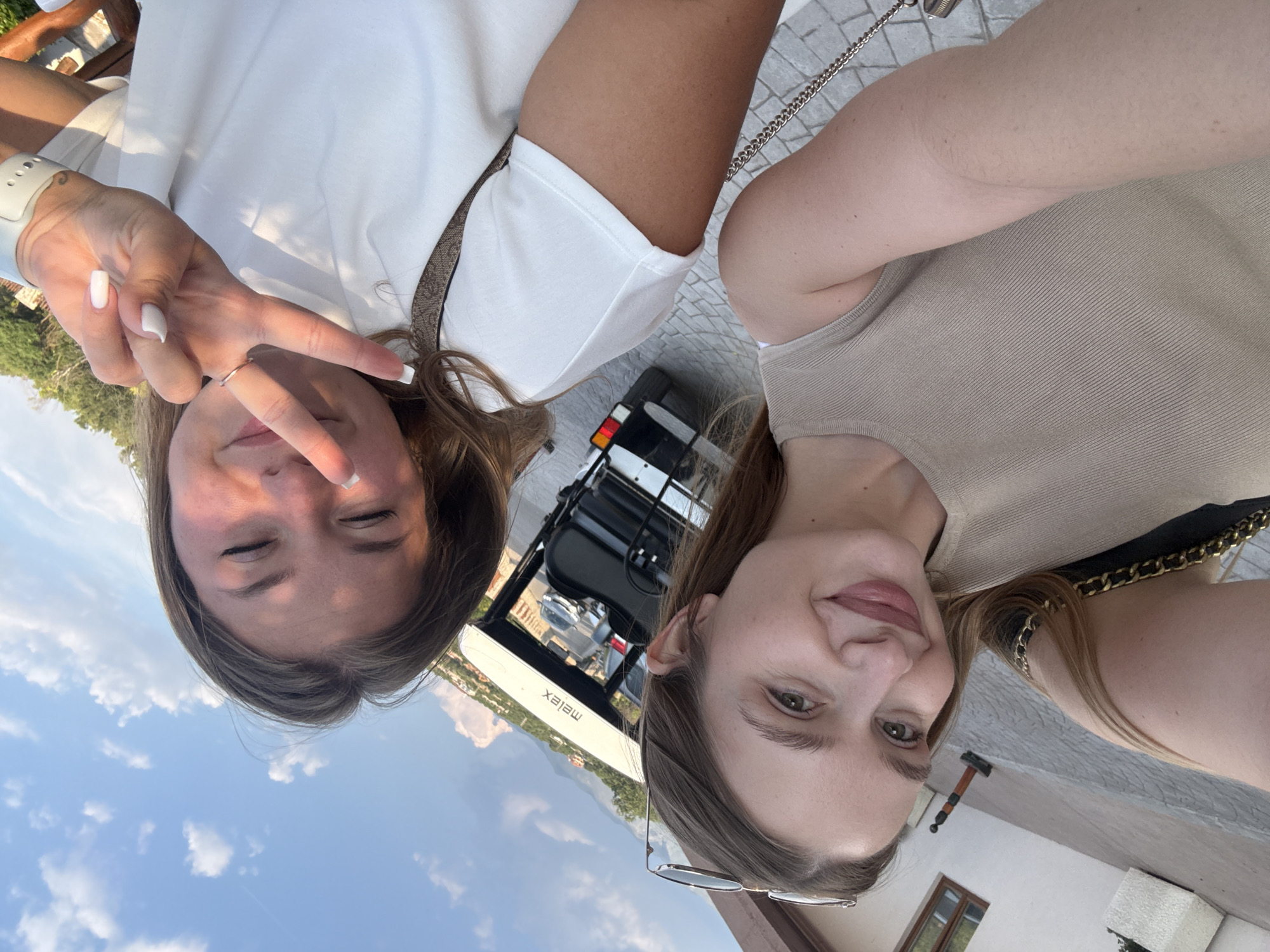 Two young women are posing for a selfie. The woman on the left is wearing a white shirt and making a peace sign with her fingers. The woman on the right is wearing a beige top and has glasses on the top of her head. Behind them is a golf cart. In the background, a mountain and clear sky are visible. The location is Kovačko Polje, Montenegro.