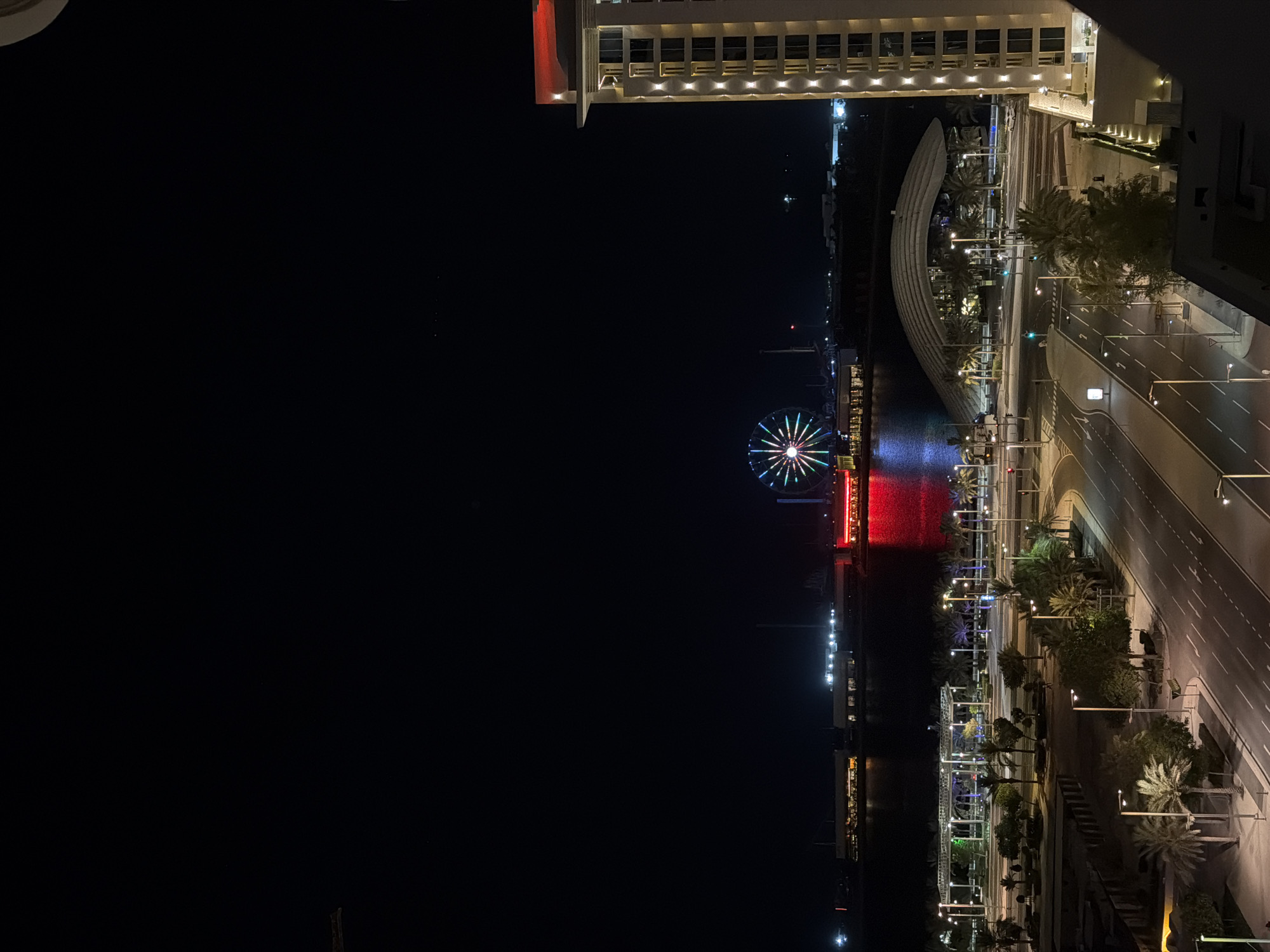 A high-angle, night-time exterior shot captures a city scene with a prominent Ferris wheel and a modern building. The Ferris wheel, illuminated with a vibrant rainbow of colors, stands in the distance on the waterfront. Adjacent to the Ferris wheel are several buildings with lights. In the foreground, a multi-lane road curves through the scene, lined with streetlights and palm trees. A sleek, modern building with illuminated windows dominates the right side of the frame, its top edge adorned with red lighting. The sky is dark, indicating nighttime.