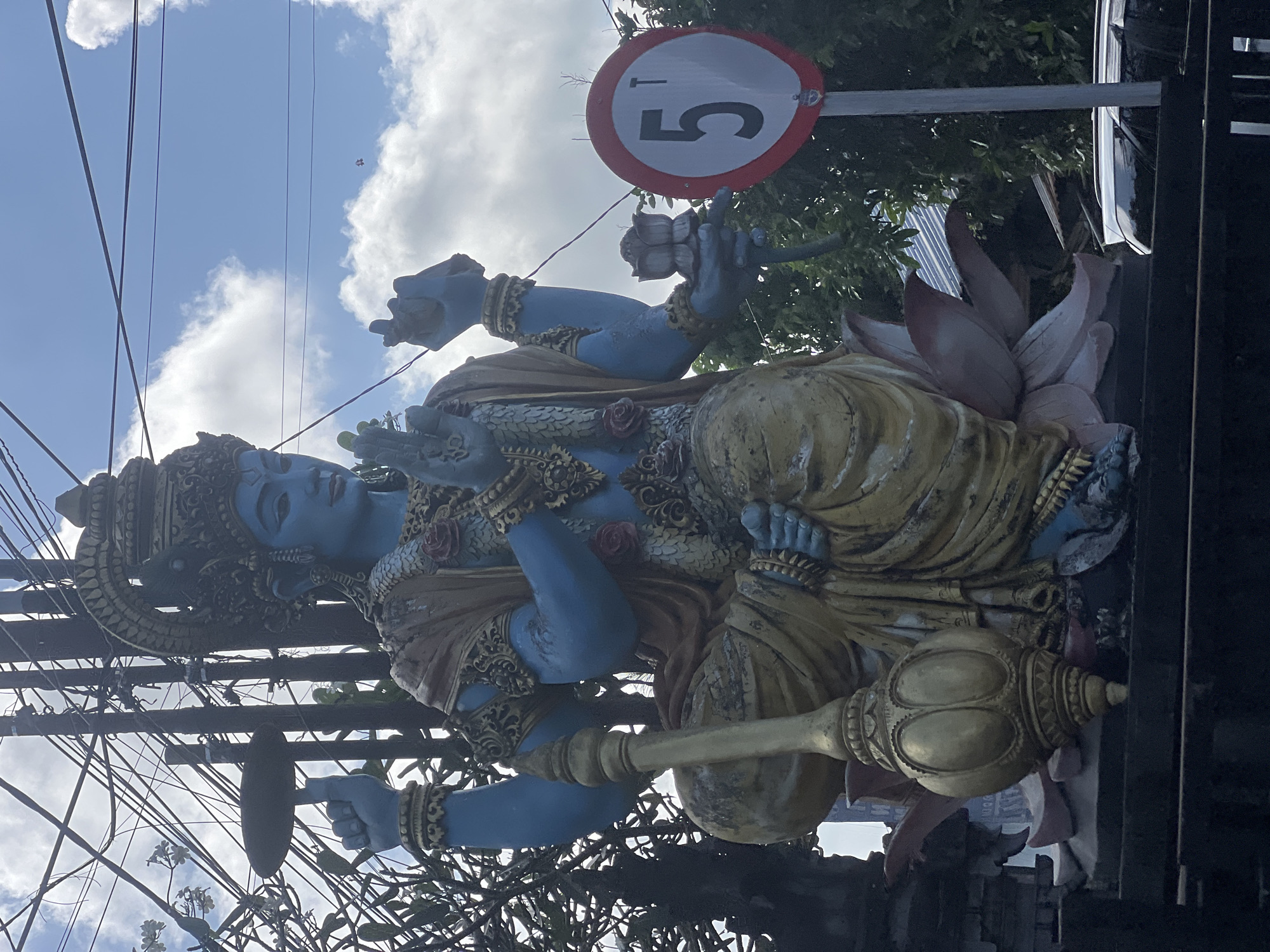 The image captures a reclining blue statue of Vishnu, adorned with golden ornaments, resting on a lotus flower pedestal. Vishnu is holding a lotus flower and a gada (mace). A speed limit sign with the number '5' is seen partially obscured behind the statue. Power lines intersect the view of the sky. The statue is located in Sukawati, Indonesia.