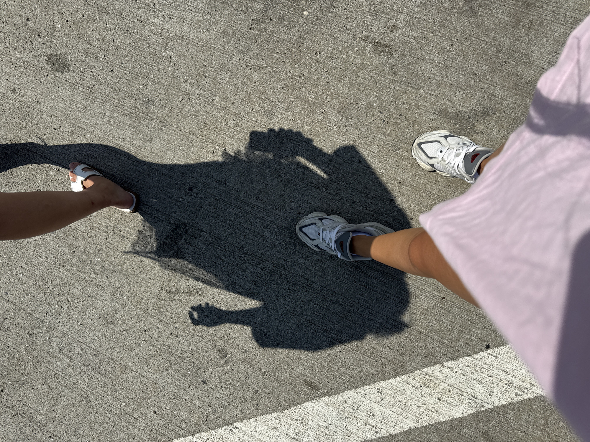 The image shows a low-angle view of two pairs of legs and their shadows on a concrete surface marked with a white line. The person in the foreground is wearing a light pink garment and white sneakers with grey and red accents. Their shadow, cast on the concrete, is large and shows a raised hand. The second person, slightly further away, is wearing a white sandal and their shadow is visible. The concrete appears to be a sidewalk or pathway. The scene is lit by bright sunlight, creating distinct shadows. The location is identified as Sanpetru, Romania.
