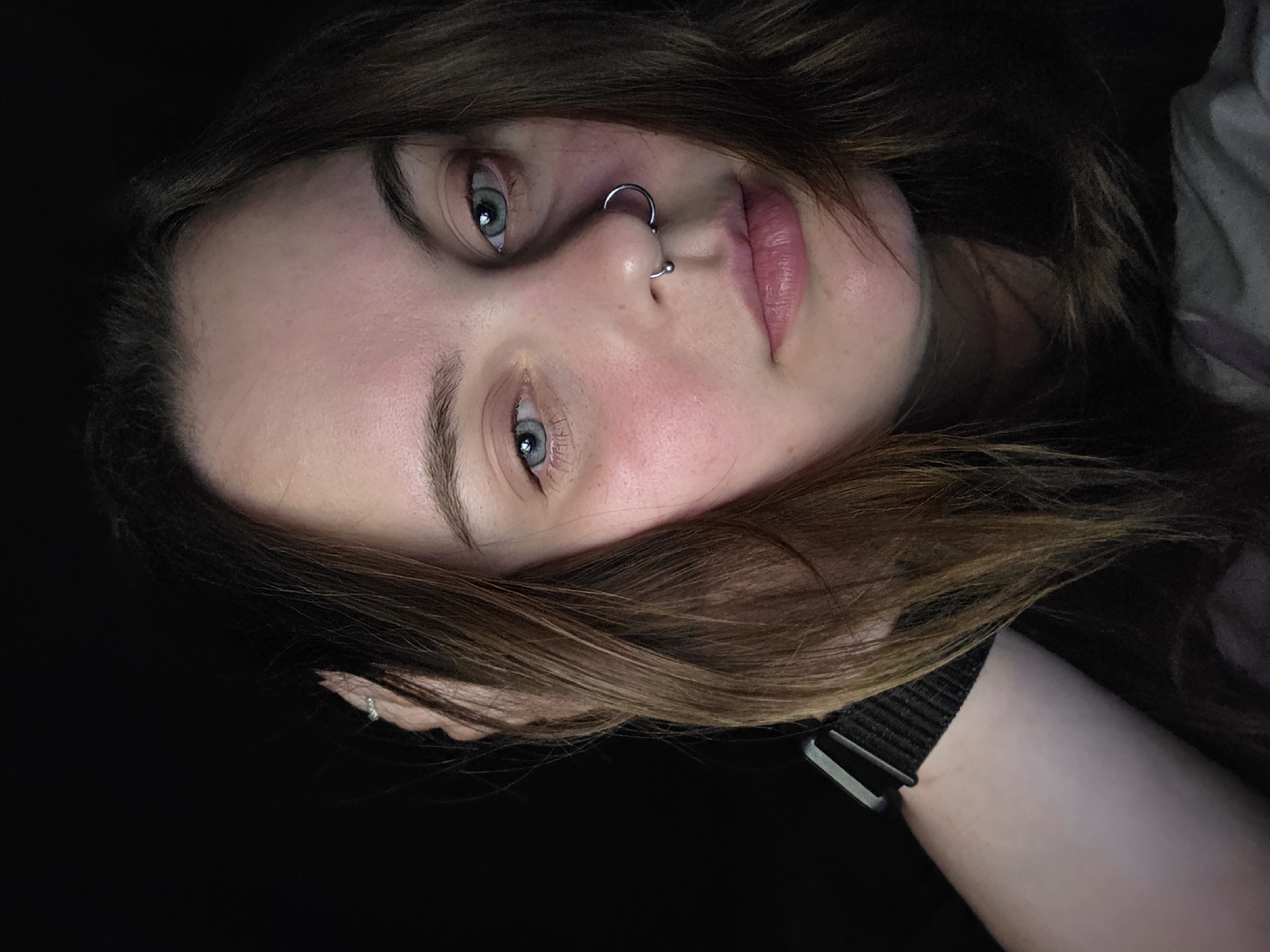 A close-up, downward-tilted portrait of a fair-skinned woman with light brown hair. She has blue eyes and a double nose piercing, consisting of two silver-colored hoops. Her lips appear pink and slightly glossy. A black watchband with a silver buckle is visible on her wrist. The background is predominantly dark, with some lighter clothing visible on the right side. The city and location are unknown.