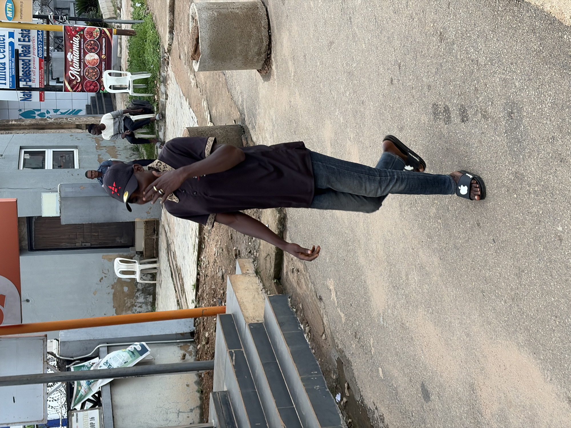 This media file captures a street scene in Jos, Nigeria. The primary subject is a young Black man standing on a paved area, possibly a sidewalk or road, in the foreground. He is wearing a dark t-shirt, blue jeans, and black slide sandals. He has a black baseball cap on his head and is looking down, with his right hand near his face.
Behind the man, a building with a light-colored facade and some signs is visible. One sign, in red and white lettering, reads "Mamamia RESTAURANT & CATERING SERVICES." Another sign, in blue and white, says "Natu-Blessed Real Esta" and contains contact information, including a phone number and a location cue "PLOT 9967 YAKUBU GOWON EXPRESS WAY, DADIN KOWIA JOS." There are also other people in the background, some sitting on chairs and others standing, suggesting a public space with commercial activity. A white plastic chair is visible near the building. The ground is paved with asphalt and some stone tiles. The overall setting appears to be an urban environment during daylight hours, with the sun casting shadows. The scene conveys a casual, everyday atmosphere.