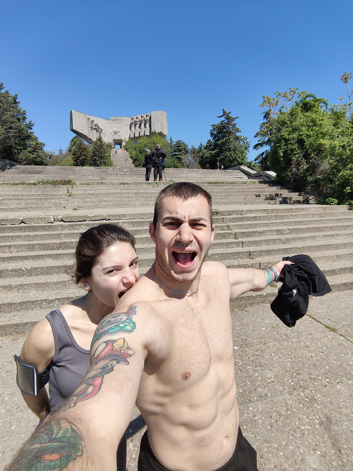 A medium shot captures a shirtless man in the foreground, his mouth open in a shout, holding a black t-shirt in his right hand. He sports a full arm tattoo on his left arm and a silver chain necklace. Behind him, a woman in a gray tank top is playfully biting his shoulder, her expression mischievous. The backdrop features a wide staircase leading up to a monumental structure, possibly a war memorial, with figures and inscriptions on its facade. Two individuals are seen standing near the base of the monument, and greenery, including trees and shrubs, frames the scene. The sky above is clear and blue. The location is indicated as Unknown, Unknown.