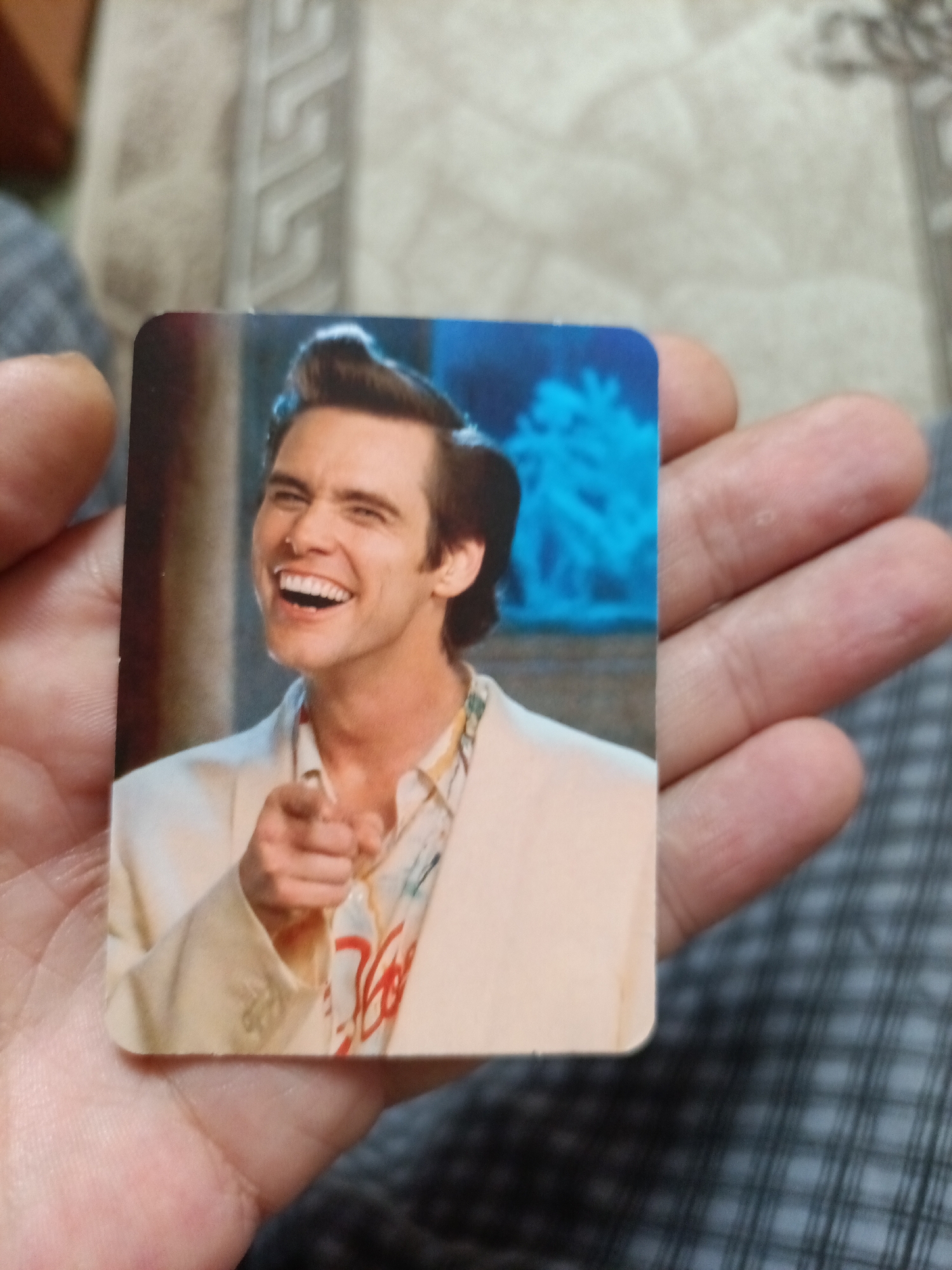 A close-up, slightly blurred, overhead shot shows a hand holding a rectangular photograph. The photograph features a still image of actor Jim Carrey in character as Lloyd Christmas from the movie "Dumb and Dumber". He is wearing a light-colored suit jacket over a loud, patterned shirt, and has a wide, toothy grin, with his head tilted slightly and his right index finger pointing forward. The background of the photograph is a blurry blue. The hand holding the photograph is light-skinned and appears to be resting on a dark, checkered fabric surface. The surrounding area of the photograph is out of focus, showing parts of a hand and a patterned textile. There is no discernible connection or relevance to the city of Nyagan, Russia.
