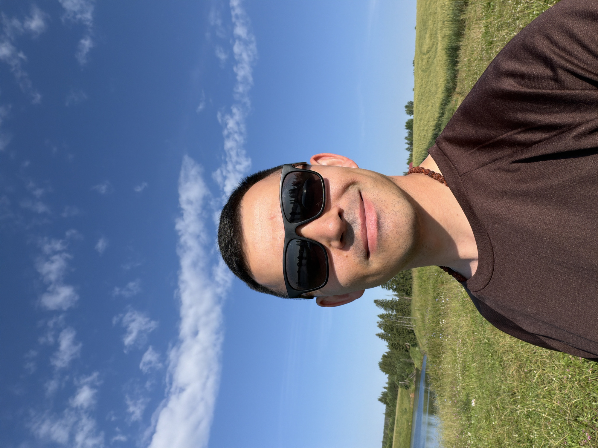 A man with short dark hair and sunglasses is taking a selfie outdoors. He is wearing a brown t-shirt and a beaded necklace. Behind him, there is a grassy field with a body of water to the left and a forest in the background. The sky is blue with some white clouds. The location is the city of Podshivalovo, Russia.