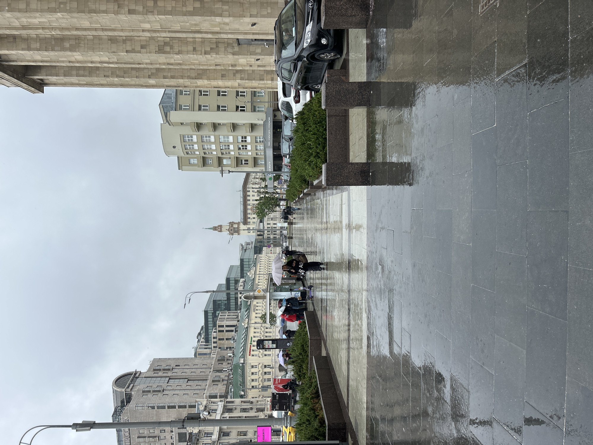 The image captures a rain-soaked city street, likely in Moscow, Russia, given the architectural style and signage. The foreground features a wide, grey-tiled sidewalk reflecting the wet conditions. Buildings line the street, varying in height and architectural design, including modern glass structures and older stone buildings. Several people are walking along the sidewalk, mostly under umbrellas, suggesting ongoing rain. Cars are parked along the street, and greenery separates the sidewalk from the road. The sky is overcast, further indicating a rainy day. A tower is visible in the distance.