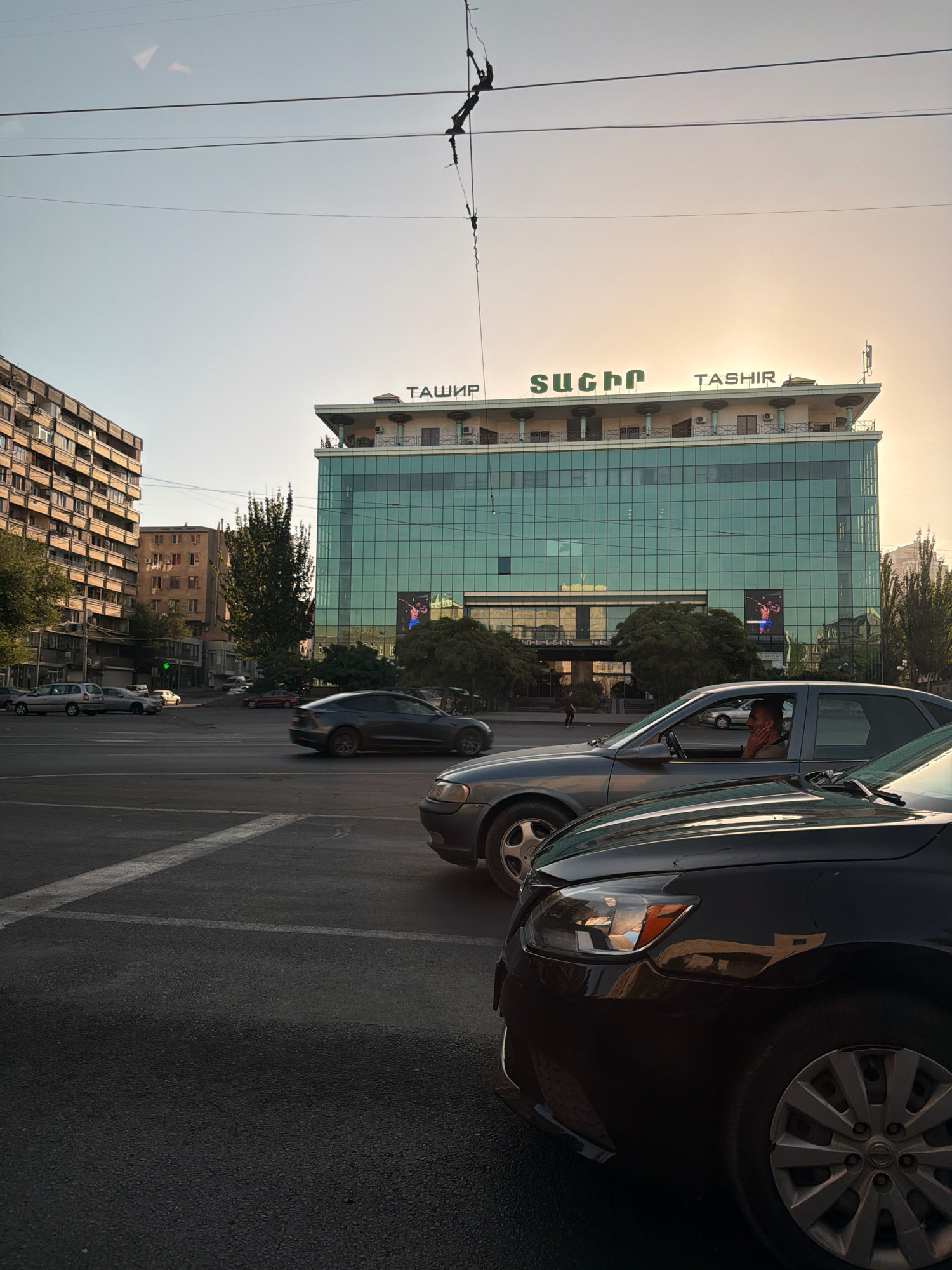 The image depicts a street scene in Yerevan, Armenia, during the late afternoon or early evening. In the foreground, a dark-colored car is visible from the front, with its headlights and wheel well in view. To its left, a gray sedan is positioned further back, with its driver visible through the window, appearing to be touching their face. Behind these cars, traffic signals and other vehicles are present on the street. The background is dominated by a modern, multi-story building with a glass facade. The building features the word "TASHIR" prominently displayed on its roof, with Cyrillic lettering also visible. Several trees are interspersed around the building and the surrounding area, and electrical wires cross the sky. The sky itself is a pale, hazy orange, suggesting either sunrise or sunset. To the left of the main building, an older, multi-story apartment building with numerous balconies is visible.