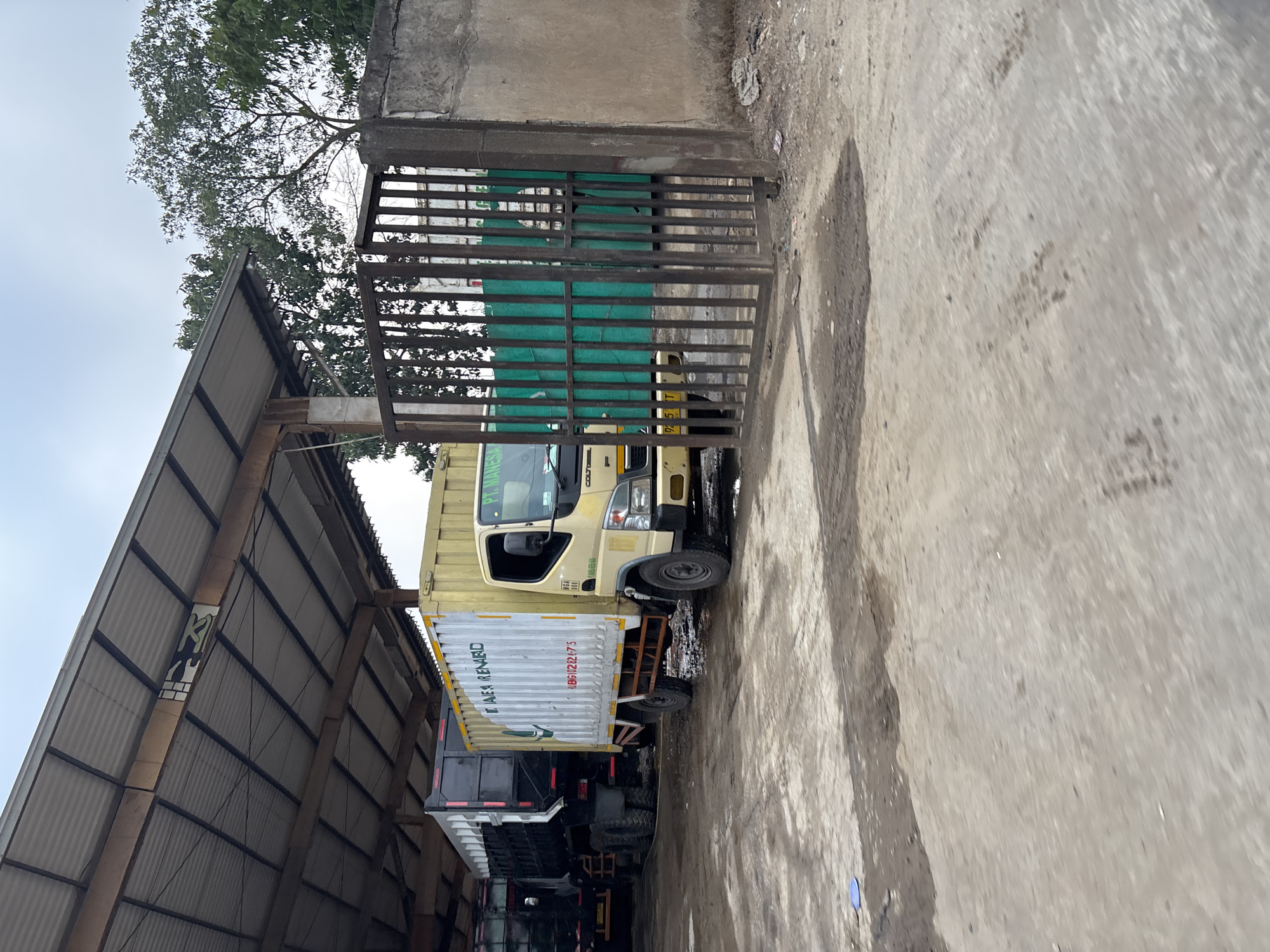 The image depicts a storage yard in Bekasi, Indonesia, under a covered open-air structure. Two trucks are parked side-by-side. The truck on the left is a black dump truck. The truck on the right is a yellow box truck with a white cargo container. The cargo container has text and logos on its side. A metal gate with vertical bars is visible in the foreground, partially obscuring the view of the trucks and the yard. Beyond the gate, a green wall or fence can be seen. The ground is paved with a concrete surface, which appears worn and somewhat dirty, with some areas of wetness or oil stains. Trees and foliage are visible in the background, suggesting an outdoor industrial or logistical area. The sky is overcast.