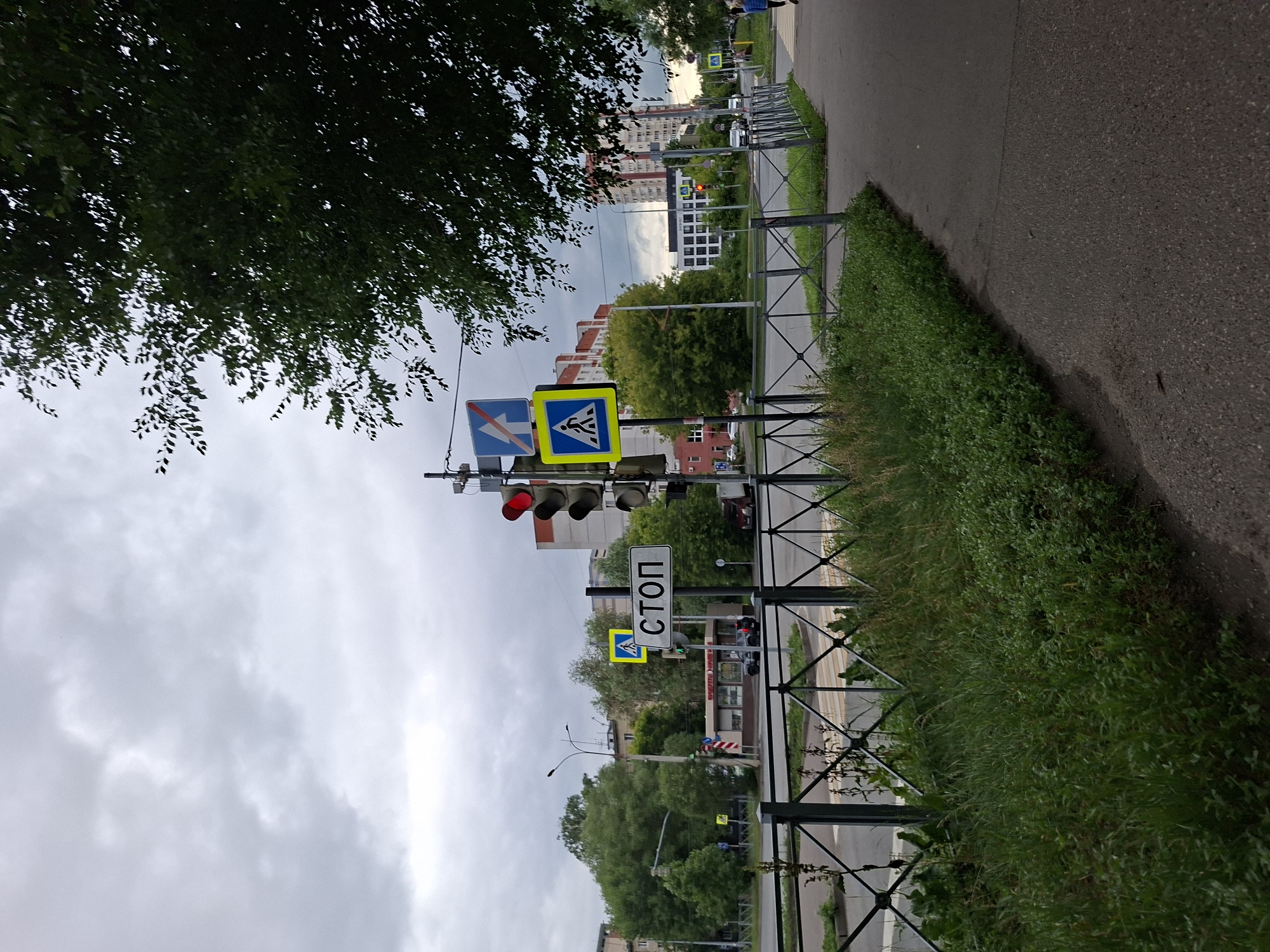The image shows a street scene in Kazan, Russia, on a cloudy day. In the foreground, there is a sidewalk with a grassy verge and a metal fence. Beyond the fence is a crosswalk and a street. Traffic lights are visible, with the red light illuminated. A "STOP" sign is posted near the traffic lights, along with pedestrian crossing signs. Several buildings are visible in the background, including apartment buildings and what appears to be a small commercial building. There are a few cars on the street. A lone figure is walking on the sidewalk in the distance. The overall impression is a typical urban intersection with traffic control measures in place.