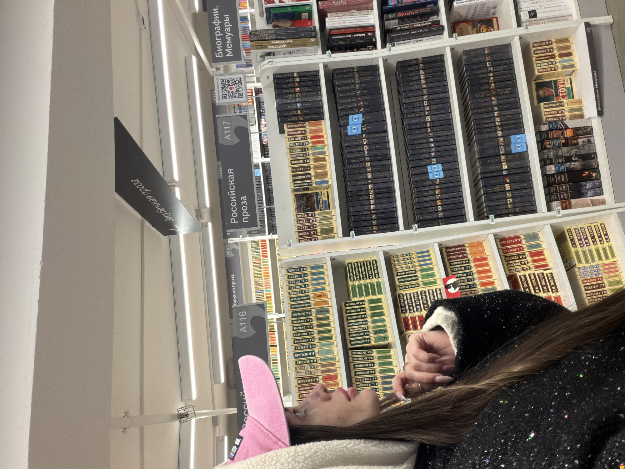 The image captures an indoor view of a bookstore or library in Saint Petersburg, Russia, showing a person browsing bookshelves. The setting is well-lit with fluorescent strip lights running parallel to the shelves.
In the foreground, a person, presumably female, is seen in profile, looking upwards at the books. She has long, dark hair and is wearing a pink baseball cap and a dark, sparkly jacket or coat with a white or cream fuzzy lining. Her hands are visible, with painted nails, resting near her face. Her expression appears calm and focused on the books.
The background is dominated by tall white bookshelves filled with numerous books, primarily in Russian, organized by author and genre. The spines of the books are mostly dark blue, black, or beige, with titles printed vertically. Shelf dividers are visible, labeled with genres and section numbers.
Visible text on the shelf labels and book spines includes: "Биографии. Мемуары" (Biographies. Memoirs), "Российская проза" (Russian prose), and "Зарубежная проза" (Foreign prose), along with section identifiers like "A117", "A116", "A046", and "A044". A QR code is also visible on one of the shelf labels near "A046" with the text "уникальный коллектор" (unique collector).
Numerous prominent Russian authors are identifiable on the book spines, such as F.M. Dostoevsky (Ф.М. ДОСТОЕВСКИЙ), L.N. Tolstoy (Л.Н. ТОЛСТОЙ), A.S. Pushkin (А.С. ПУШКИН), A.P. Chekhov (А.П. ЧЕХОВ), Gogol (ГОГОЛЬ), M.A. Sholokhov (М.А. ШОЛОХОВ), and Maxim Gorky (МАКСИМ ГОРЬКИЙ). Other visible names include Vladimir Vysotsky (ВЛАДИМИР ВЫСОЦКИЙ), I.A. Goncharov (И.А. ГОНЧАРОВ), and Bulgakov (БУЛГАКОВ). Titles like "БЕСЫ" (Demons, by Dostoevsky), "ОБРЫВ" (The Precipice, by Goncharov), and "НА ДНЕ" (The Lower Depths, by Gorky) are also readable. Some non-Russian titles or subjects like "ПИКАССО, ИНОСТРАНЕЦ" (Picasso, Foreigner) and "50 ПРАВИЛ КОКО ШАНЕЛЬ" (50 Rules of Coco Chanel) are also visible in the "Биографии. Мемуары" section.