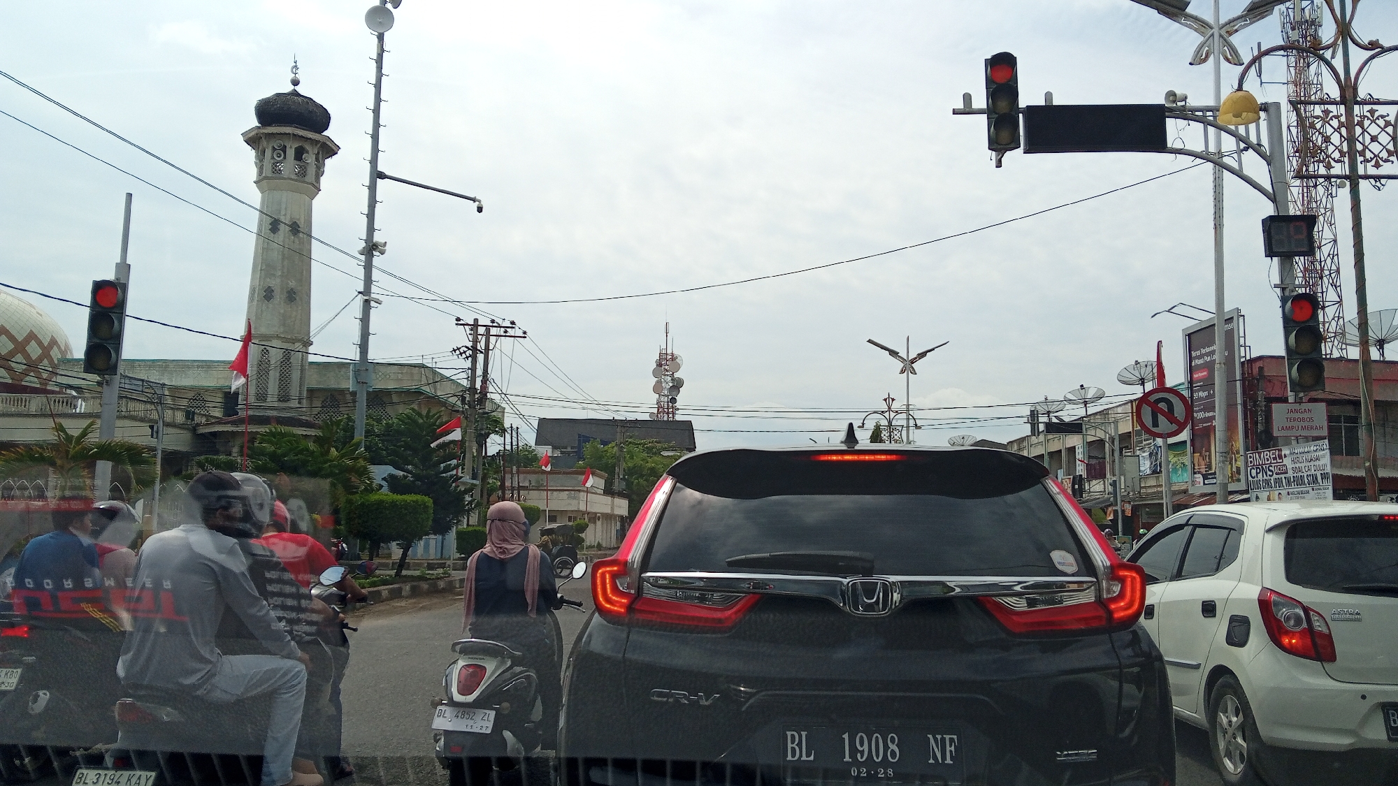 The image shows a traffic scene in Lhokseumawe, Indonesia, with a mosque and its minaret visible on the left side. A traffic light with a red signal is displayed. Several vehicles are present, including a dark-colored Honda CR-V in the foreground, a white car behind it, and multiple motorcycles. People on the motorcycles are wearing helmets and one person is wearing a headscarf. Buildings, utility poles with wires, and antennas are in the background. The sky is overcast.