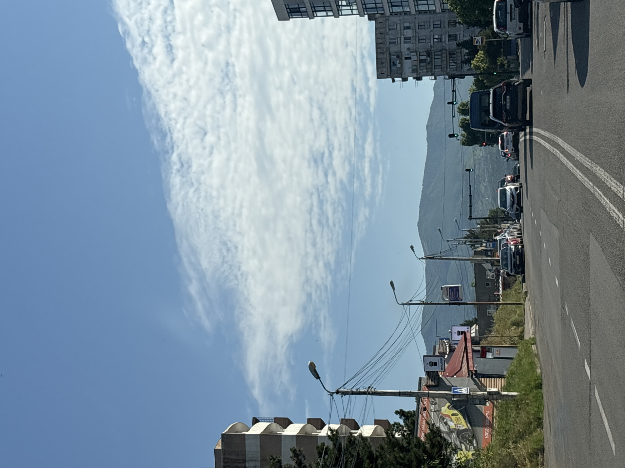 The image captures a street scene in Tbilisi, Georgia, on a clear day with scattered clouds in a blue sky. The foreground features a two-lane road with white lane markings, indicating traffic flow. Several vehicles are visible on the road, including cars and a van, suggesting moderate traffic.
On the left side of the road, a multi-story building with curved balconies stands prominently, partially obscured by trees. Adjacent to it, there are smaller structures, including what appears to be a shop or cafe with signage and a large advertisement featuring a yellow sports car. Streetlights and utility poles with a complex network of wires line the street.
In the mid-ground and background, more buildings of varying architectural styles are present, some appearing to be residential apartment blocks. A range of mountains forms the distant backdrop, their slopes covered in vegetation. The presence of traffic lights and road signs indicates this is an urban or semi-urban environment within Tbilisi. No people are clearly discernible in the scene. The overall impression is that of a typical day in a city with a blend of modern and older infrastructure, set against a natural mountain landscape.