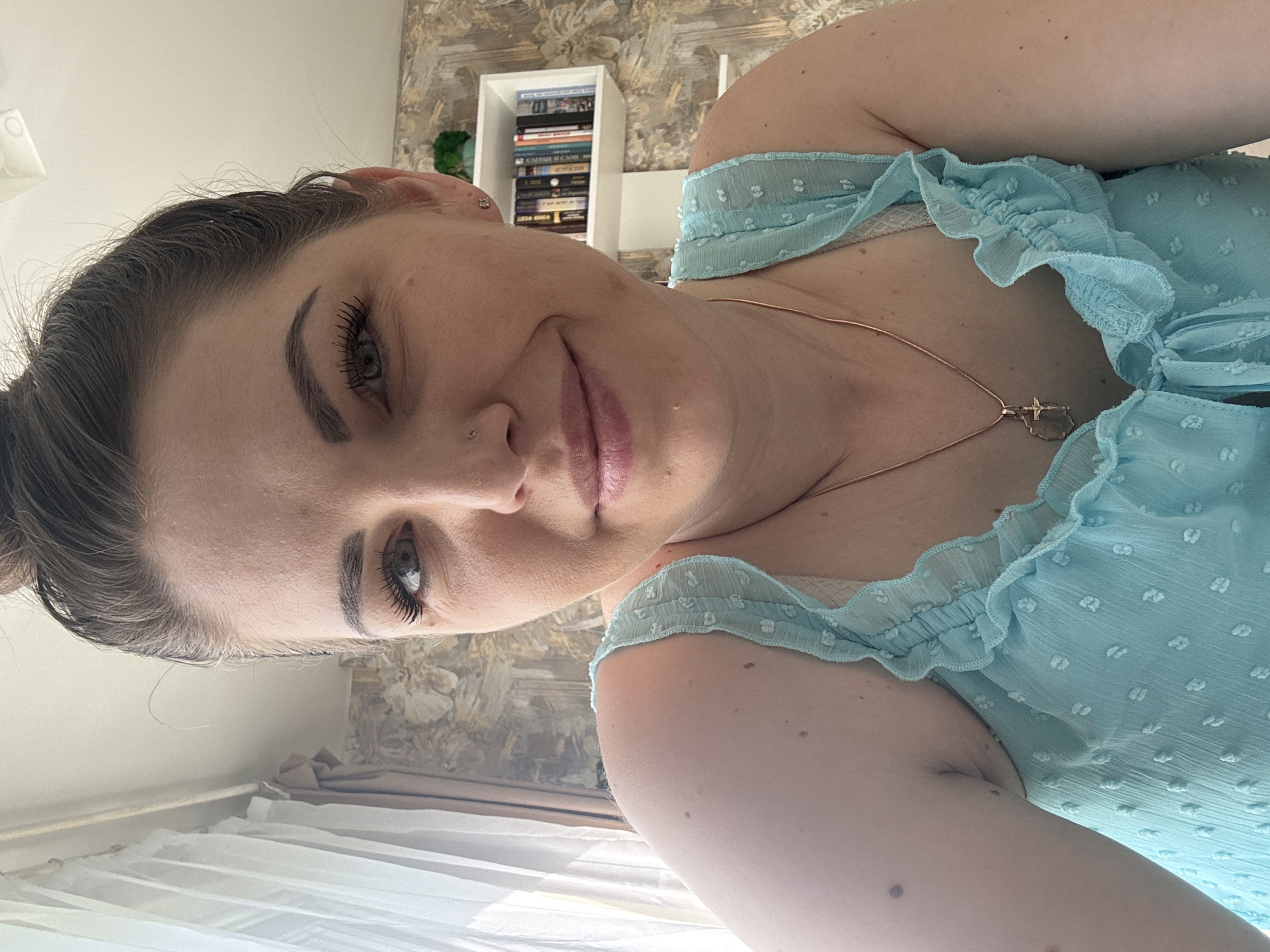 The image is a selfie of a woman with light skin and dark hair pulled back. She has prominent eyebrows, long eyelashes, and a small piercing in her nose. She is wearing a light blue, ruffled top and a thin gold necklace with a pendant. In the background, there is a bookshelf filled with books and a patterned wall. A window with white curtains is visible in the lower left corner. The city of Razdolie, Russia, cannot be inferred from the image.