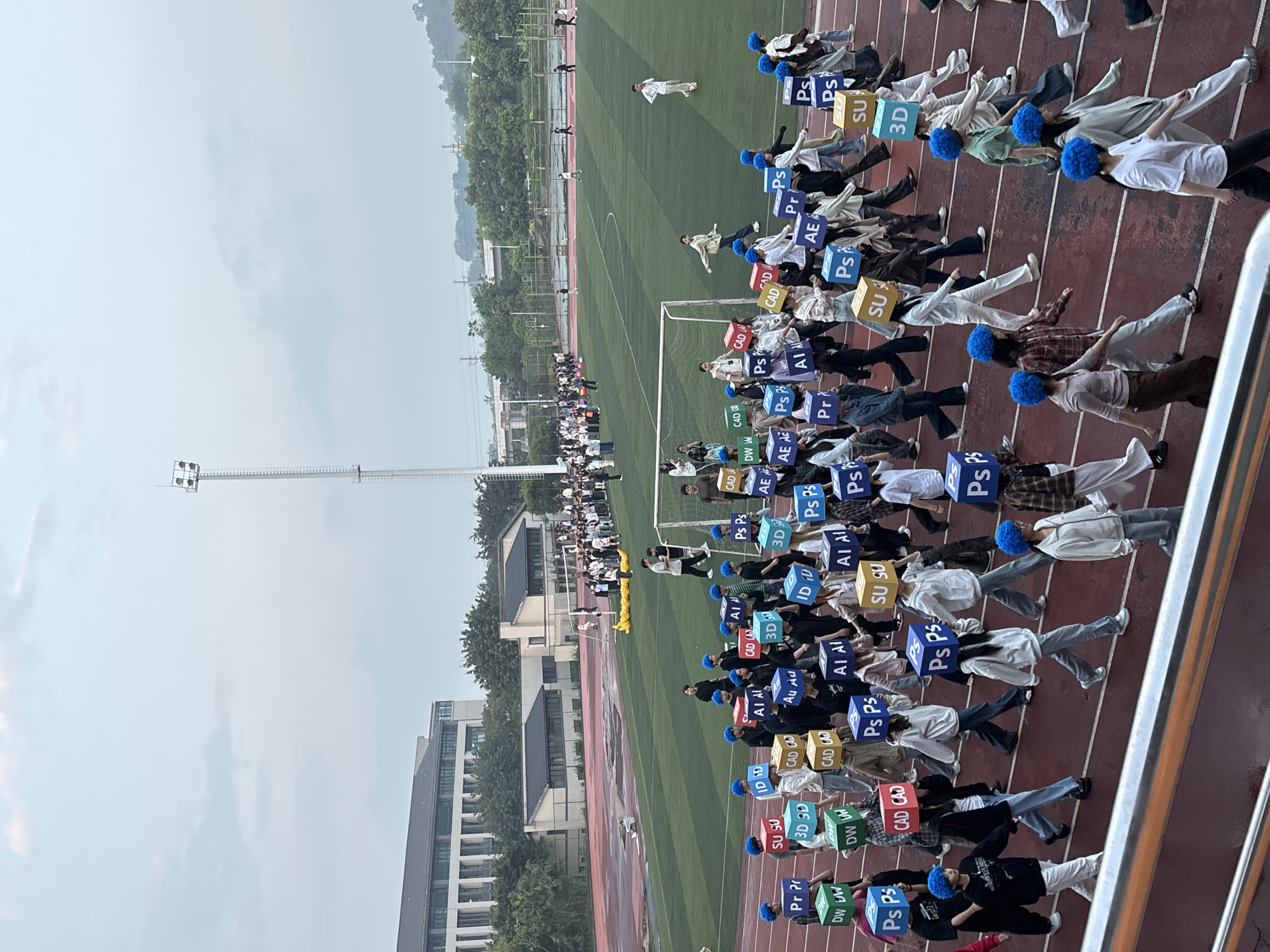 Here is a detailed description of the image:
**Scene:** A wide-angle, ground-level shot of a stadium at a university or school in Jiangjin, China. The stadium features a green field, surrounded by a running track with lanes marked in red-brown. A large group of students are on the track, likely participating in a demonstration or choreographed event.
**People:** The students are mostly dressed casually in light-colored tops and jeans, and many are wearing blue hats or wigs. Each student appears to be holding a square sign displaying an abbreviation, such as "PS," "3D," "CAD," "AI," "DW," "SU," "AE," and "Pr".
**Objects:** Key objects include:
* Square signs bearing abbreviations.
* A soccer goal.
* Running track.
* Lanes.
* Field.
* School buildings.
* Light tower.
* Trees.
* Sidewalk.
* Stadium.
**Actions:** The students are aligned in rows on the running track, with the intention of creating a coordinated display using their signs. Some students are in motion.
**Environment:** The overall environment shows a stadium setting on an overcast day. The university or school buildings are visible on one side, with surrounding trees.
