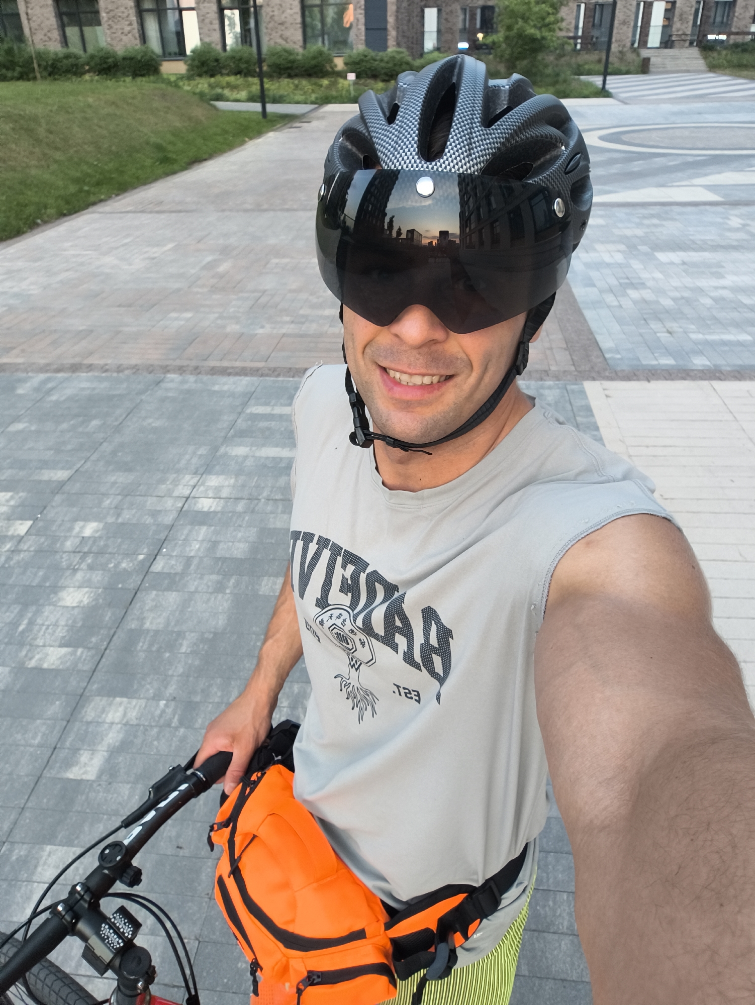 A man wearing a black bicycle helmet with a visor, a grey t-shirt, and an orange fanny pack is taking a selfie. He is holding the handlebars of a bicycle. The background shows a paved area with modern brick buildings and grassy areas. The reflection in the visor shows buildings. The location is plausibly the city of Moscow, Russia, based on the architecture and urban design.