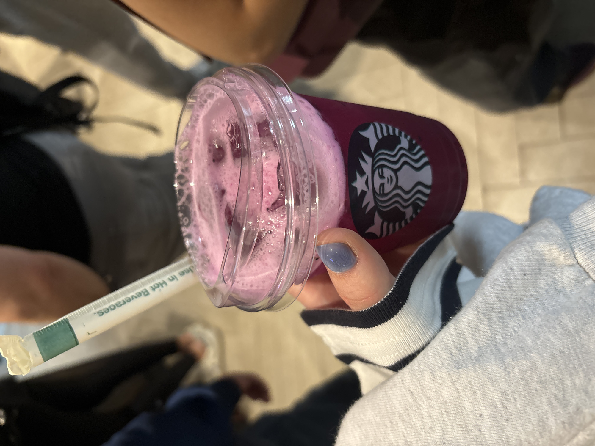 A person is holding a partially filled Starbucks drink, likely a Dragonfruit Refresher, in their right hand. The cup is made of clear plastic with a Starbucks logo on the red body. The drink inside is a pink liquid with chunks of dragonfruit. A white paper straw with green stripes is attached to the side of the cup. The person is wearing a light grey long-sleeved shirt with a striped wristband. Their fingernails are painted a light blue color. The person is standing indoors on a beige tile floor, likely in Pendik, Turkey. The background shows parts of other people's clothing.