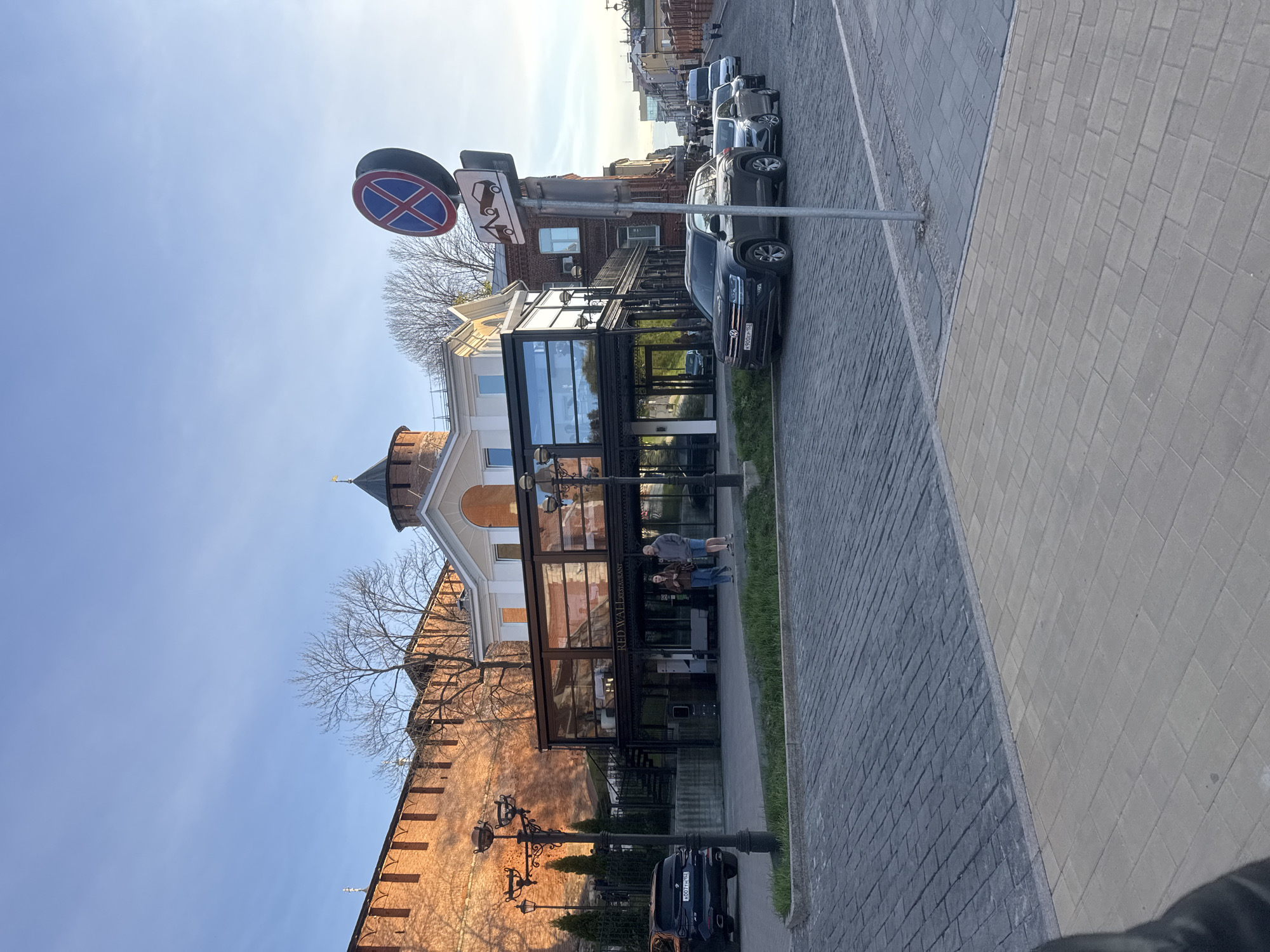 The image depicts a street in Nizhny Novgorod, Russia, featuring a blend of historic architecture and modern amenities. A segment of a brick wall with a turret is visible on the left, adjacent to a contemporary building with glass walls, housing a restaurant called "RED WALL RESTAURANT." Two people are standing on the sidewalk next to the building. Street lights are mounted along the wall. Cars are parked along the side of the road. Traffic signs are mounted on a pole. The sky is blue, and the street is paved with blocks.