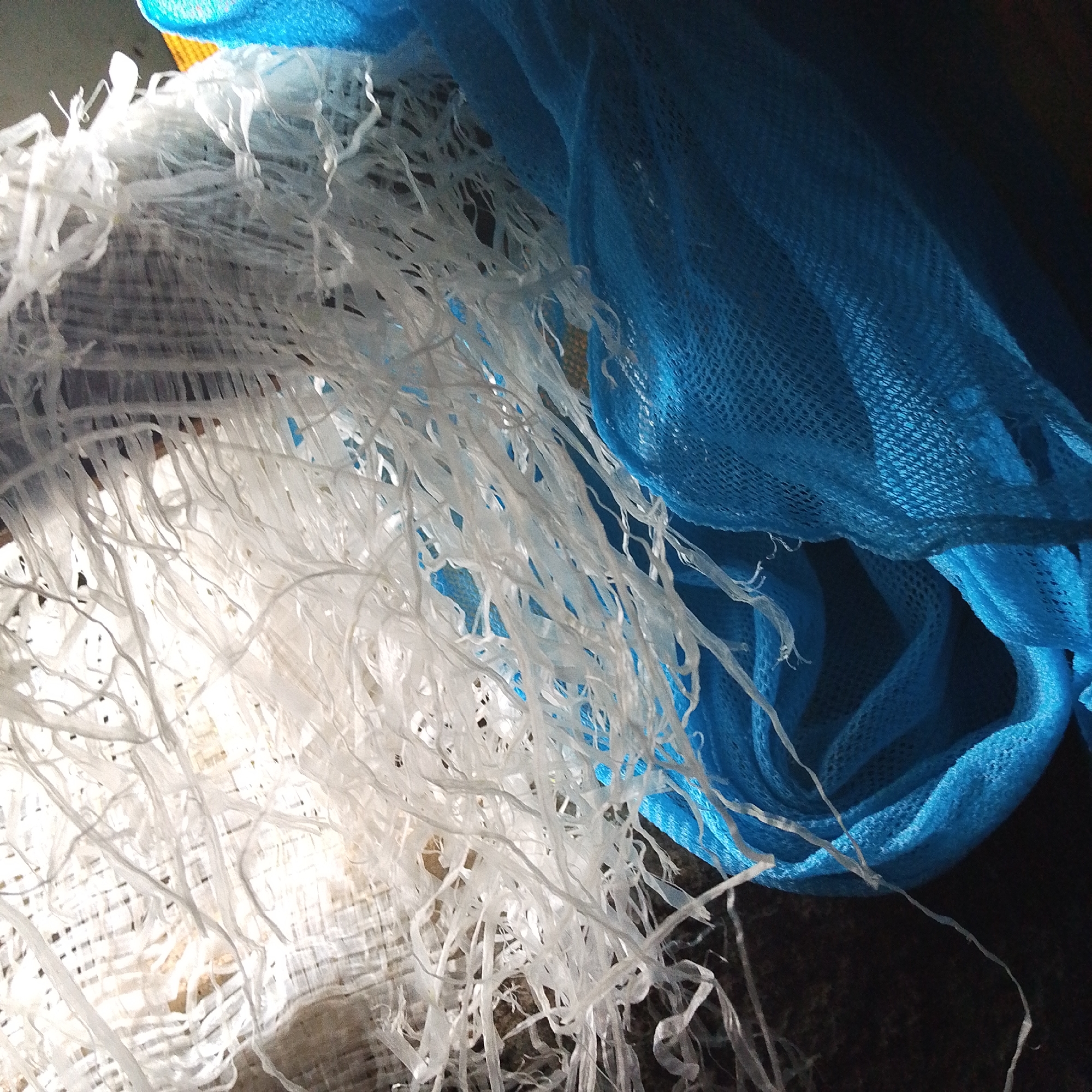 The image shows a close-up of white plastic strips tangled with a blue net-like material. The lighting suggests it is indoors, possibly in a dimly lit storage area or on a dark surface. There is no clear indication of the city of Jalingo, Nigeria, or any specific environmental context that could place it there. The scene is static and focuses on the textures and arrangement of the materials.