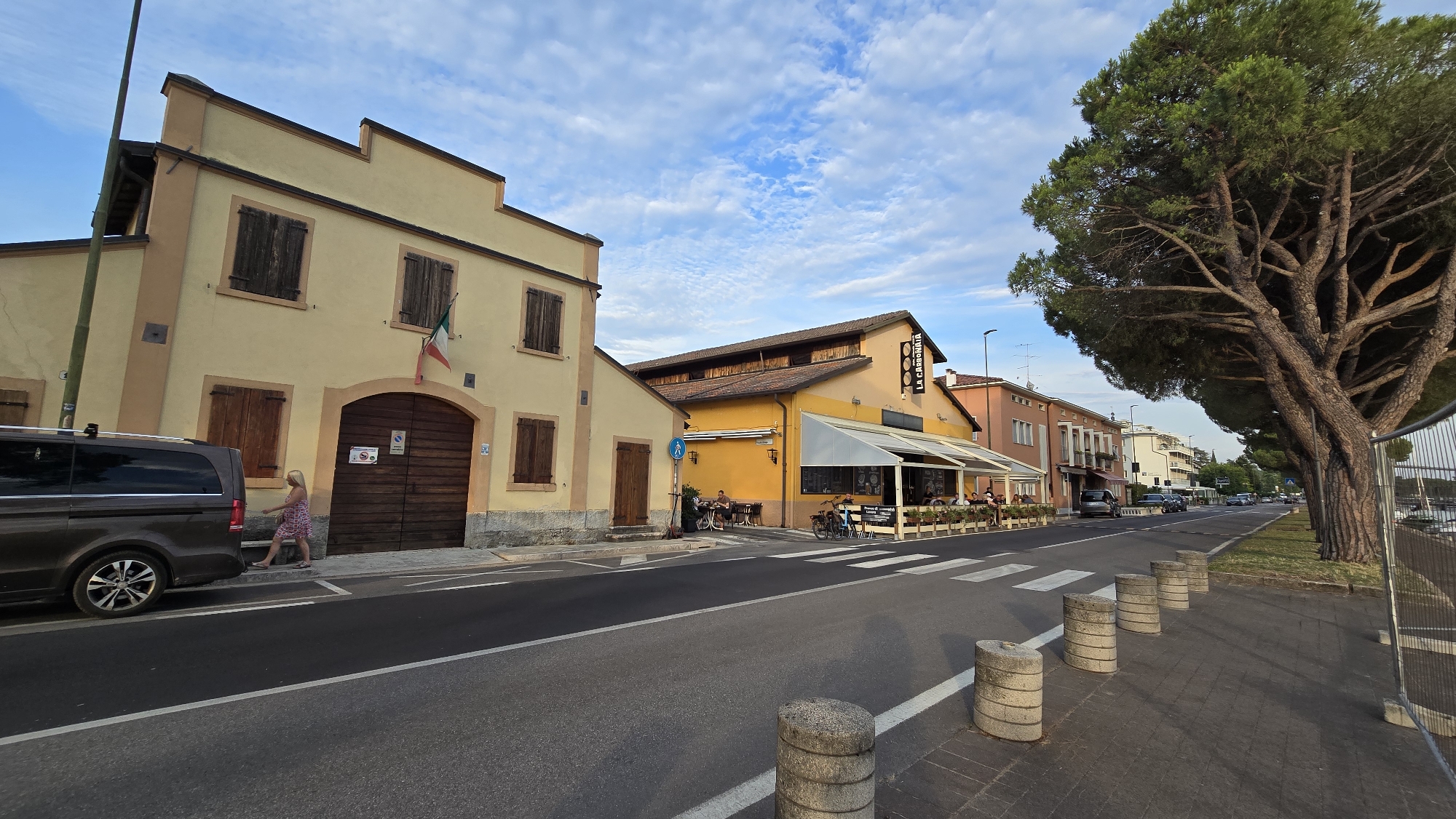 The image depicts a street scene in Peschiera del Garda, Italy, likely during the late afternoon or early evening due to the warm lighting and long shadows. The sky is partly cloudy, suggesting fair weather. The scene features a row of buildings with a mix of residential and commercial spaces.
On the left, a light yellow building with a large wooden double door and dark shutters is prominent. An Italian flag is affixed to the building. To its right, a dark brown minivan is parked. A woman in a patterned dress is walking on the sidewalk next to the minivan.
Further down the street, a yellow building with a sign reading "LA CARROZZIA" is visible, indicating a restaurant or café with outdoor seating. Several patrons are seated at tables under a white awning. Bicycles are parked nearby. The street is paved with asphalt and has a pedestrian crossing.
The right side of the image is dominated by a large, mature pine tree with a thick trunk and sprawling branches. A sidewalk runs alongside the street, lined with circular concrete bollards. A metal fence is visible along the far right edge of the image.
Cars are seen driving or parked along the street, suggesting a moderately active urban environment. The overall atmosphere appears relaxed and casual, typical of a European town in the evening. No specific event is clearly taking place, but the presence of diners and pedestrians suggests normal daily activity. The location is identified as Peschiera del Garda, Italy.