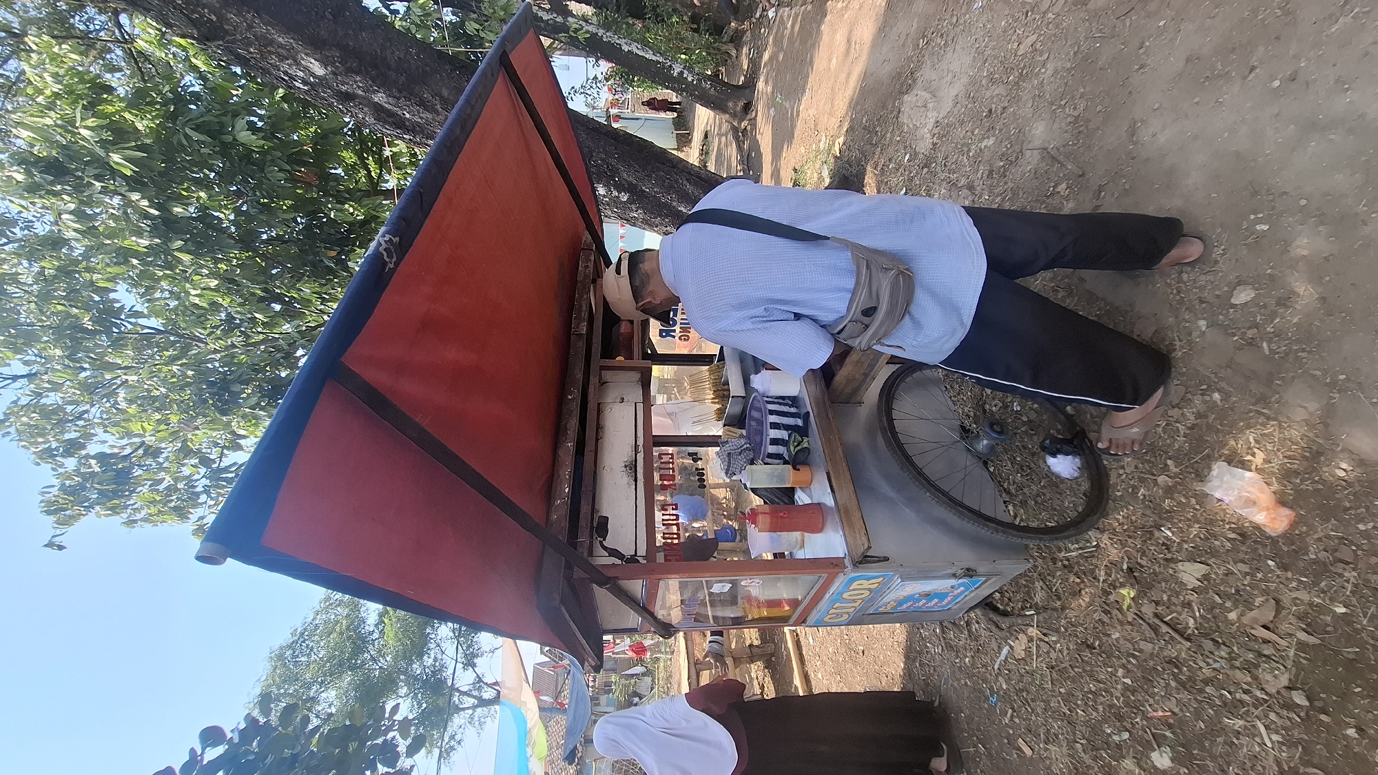 A food cart vendor, a man wearing a blue checkered shirt, black pants, and a beige baseball cap, stands at his mobile food cart under a red awning. He has a gray crossbody bag slung over his shoulder. The cart is positioned outdoors on a dirt path, with a large tree to the right and vegetation in the background. A woman in a brown dress and white hijab stands to the left of the cart, facing it. The cart has various bottles of condiments, a basket, and what appears to be skewers of food visible. A wheel is attached to the side of the cart. The overall setting suggests a street-side food vendor operation in Cilaku, Indonesia.