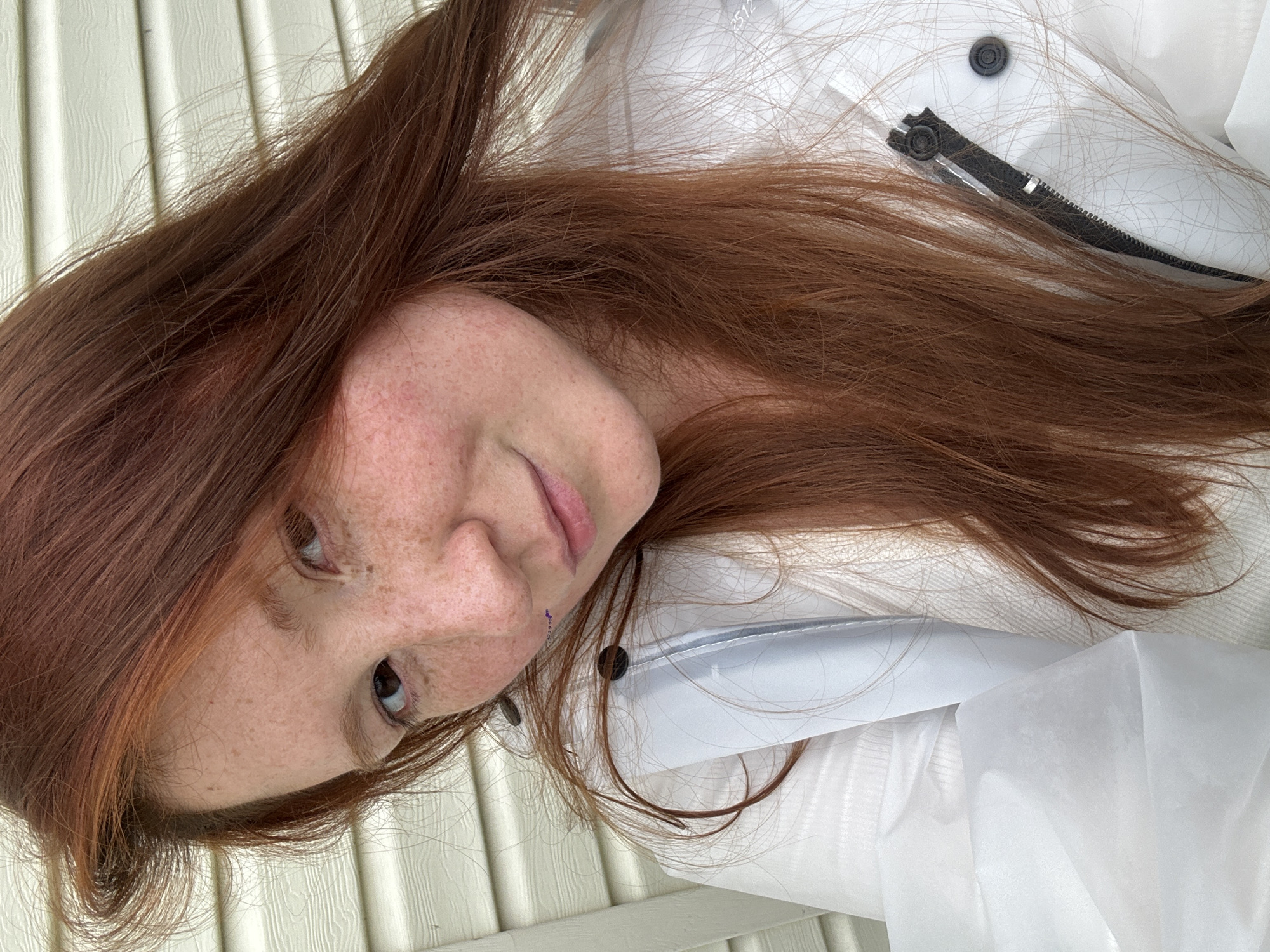 A woman with auburn hair and freckles is the central subject of the image. She is wearing a white jacket or coat with a dark-colored zipper and buttons. The background appears to be a textured, light-colored surface, possibly a wall or paneling. The scene is close-up, focusing on the woman's face and upper body.
The location cannot be determined as Nizhny Novgorod, Russia without additional visual cues or contextual information.