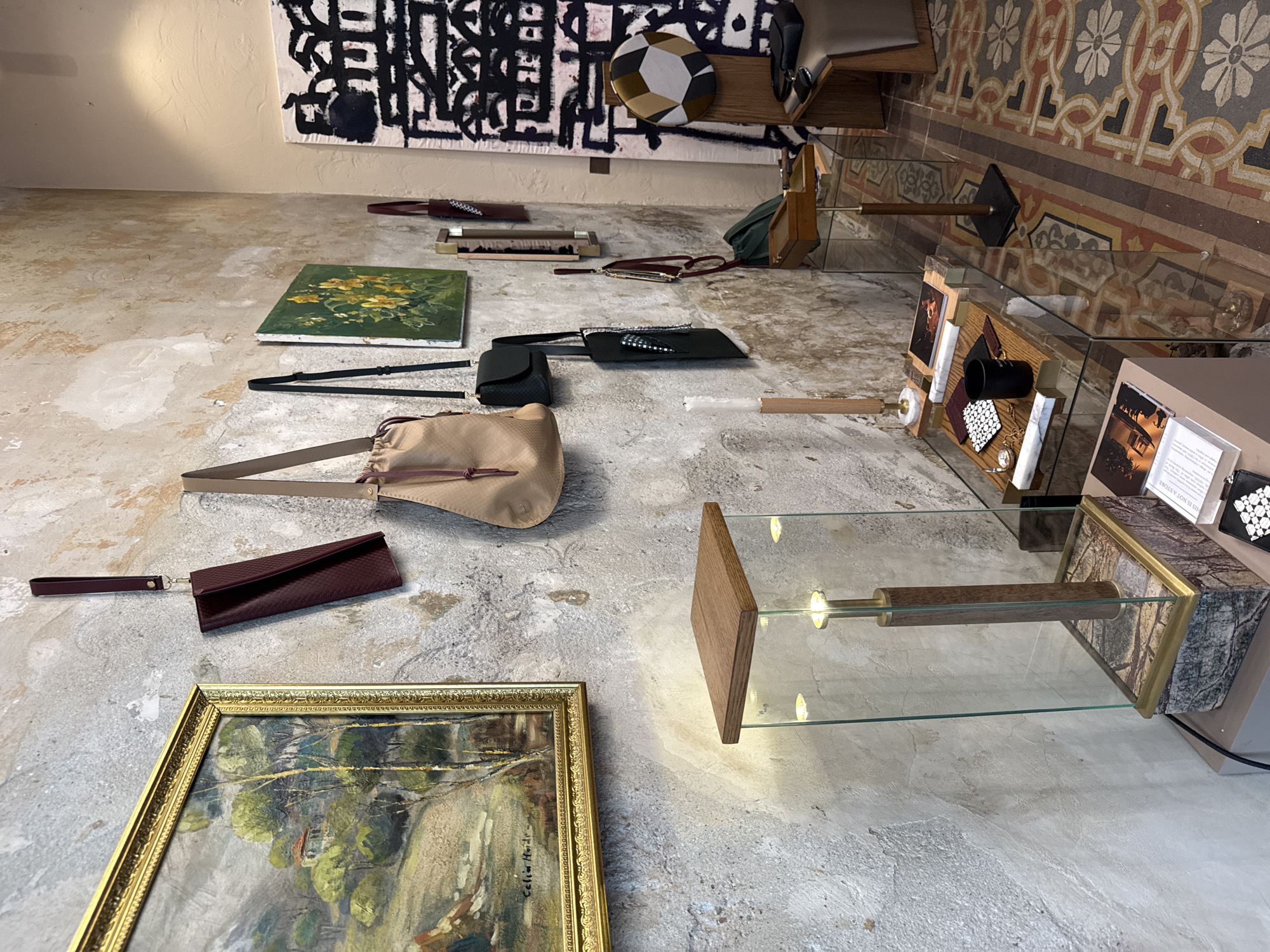 This media file captures an interior scene, likely an exhibition or boutique space in Milan, characterized by a curated display of fashion accessories, art, and decorative objects. The setting combines rustic architectural elements with modern and traditional items.
The foreground and midground feature various handbags and clutches laid out on a rough, light-colored floor. Prominently displayed are a beige drawstring bag and a burgundy clutch with a wrist strap. Several black handbags and clutches are also visible. Two paintings are placed on the floor: a small square canvas depicting yellow flowers on a green background, and a larger landscape painting in a gold ornate frame, signed "Celine Houl..." along the bottom edge.
To the right, a series of glass display cases, constructed with wooden and possibly marble bases, showcase smaller items. These cases are internally lit, illuminating a black cup, a white and black patterned tie or fabric, and a gold watch among other less discernible objects. A small sign inside one case reads, "THIS IS NOT A STORE."
The background consists of a light-colored, textured wall adorned with a large, abstract black and white patterned rug. Above one of the display cases, a circular decorative object with gold, white, and black geometric patterns is placed on a wooden shelf. Towards the far right, traditional ornate floor tiles in shades of orange, grey, and black are visible, hinting at the historical architecture of the location. No people are present, and the artificial lighting indicates an indoor setting.