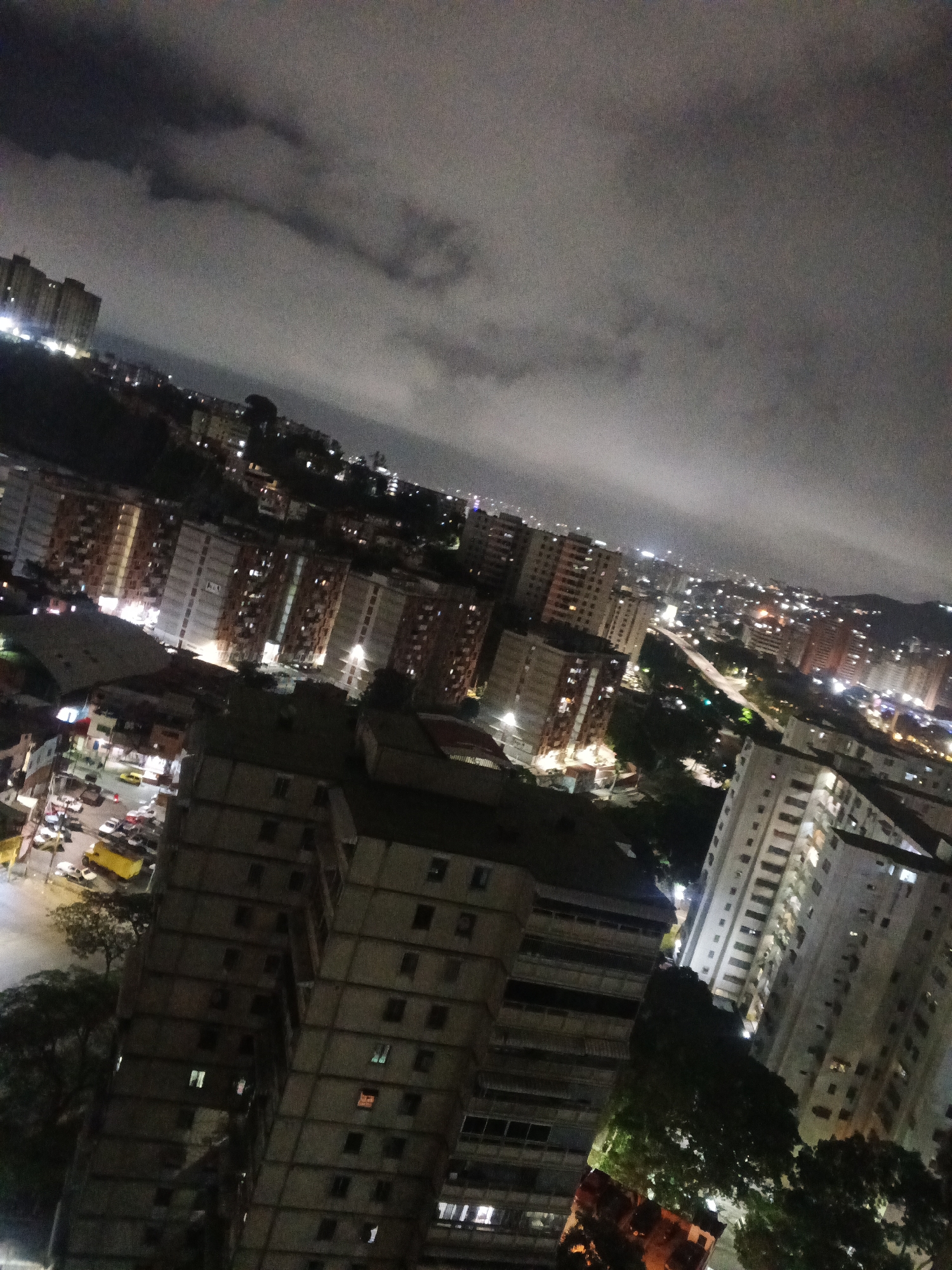 The image depicts an elevated nighttime view of Caracas, Venezuela, under a cloudy sky. Tall buildings dominate the foreground and middle ground, their windows showing scattered illumination. In the lower left, a street intersection is visible with parked and moving vehicles. Further in the distance, city lights stretch across the landscape, rising up the hills. The general atmosphere is dimly lit.