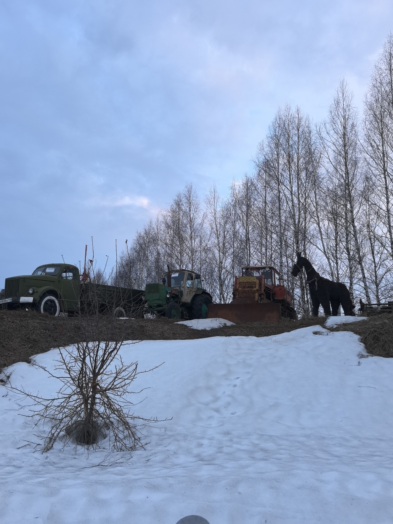 The image depicts a snowy landscape in Bol'shoi Mashlyak, Russia, with several vehicles and a horse statue positioned on a low hill against a backdrop of bare trees and a cloudy sky. From left to right, there is a green truck with a flatbed, followed by a green tractor and a red bulldozer. Next to the bulldozer stands a dark-colored horse statue. In the foreground, there is a small, leafless shrub protruding from the snow-covered ground. The overall scene is suggestive of a rural or semi-rural environment.