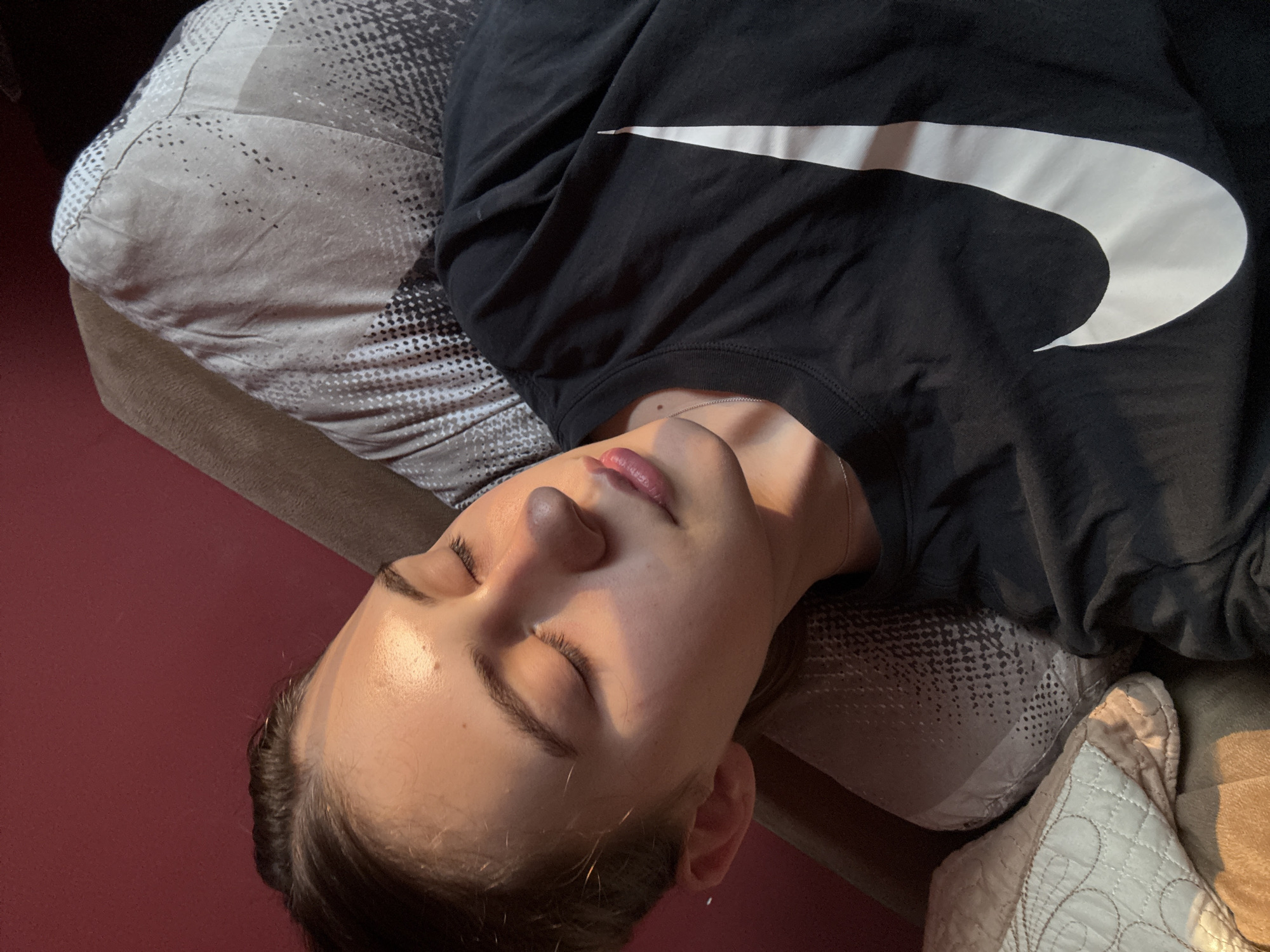 A young female with dark hair pulled back is asleep, lying on her right side, with her head resting on a patterned pillow. She is wearing a black t-shirt with a large white Nike logo across the chest. The background is a reddish-pink wall and other pillows. The lighting suggests it is indoors, with sunlight streaming in from the left, casting a warm glow on her face. There are no specific elements in the image that can be used to determine a location within Panagyurishte, Bulgaria.