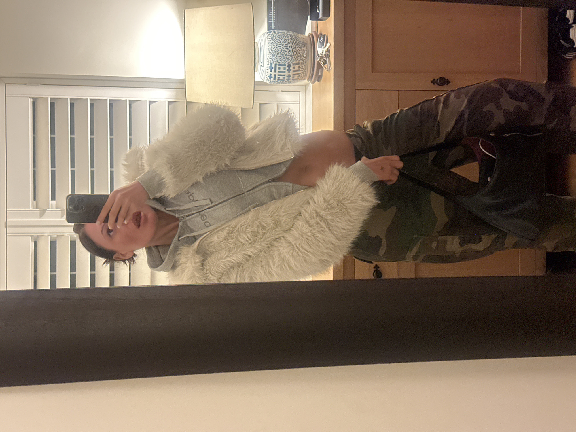 An adult female is depicted in a mirror selfie within an indoor domestic setting. She holds a dark-colored smartphone, partially obscuring her face from the upper nose downwards, as her lips are slightly parted. Her dark hair is pulled back. She wears a cream-colored faux fur jacket over a light grey cropped zippered hoodie, the latter displaying a reversed light-colored "bebe" logo on the chest and exposing her midriff. Her lower body is clad in dark green camouflage-patterned cargo pants. In her left hand, a black shoulder bag containing a maroon item is held; a small, dark tattoo is visible on the back of this hand.
The reflected background features a window fitted with white horizontal plantation shutters. Beneath the window is a light-toned wooden dresser or cabinet, equipped with two visible doors and dark-colored pulls on the right side. Situated on the dresser is a table lamp with a cream-colored fabric shade and a decorative blue and white ceramic base, resting on a dark wooden stand. A set of keys on a ring and a dark rectangular object are positioned on the dresser surface to the right of the lamp. A portion of a black electronic device is also visible further to the right. The floor beneath the dresser is a light wood or laminate, with black and white striped footwear partially visible on the far right. The mirror's thick, dark brown frame is present on the left, next to a plain light-colored wall. The specific location within Tunbridge Wells, United Kingdom, cannot be determined beyond it being an interior.