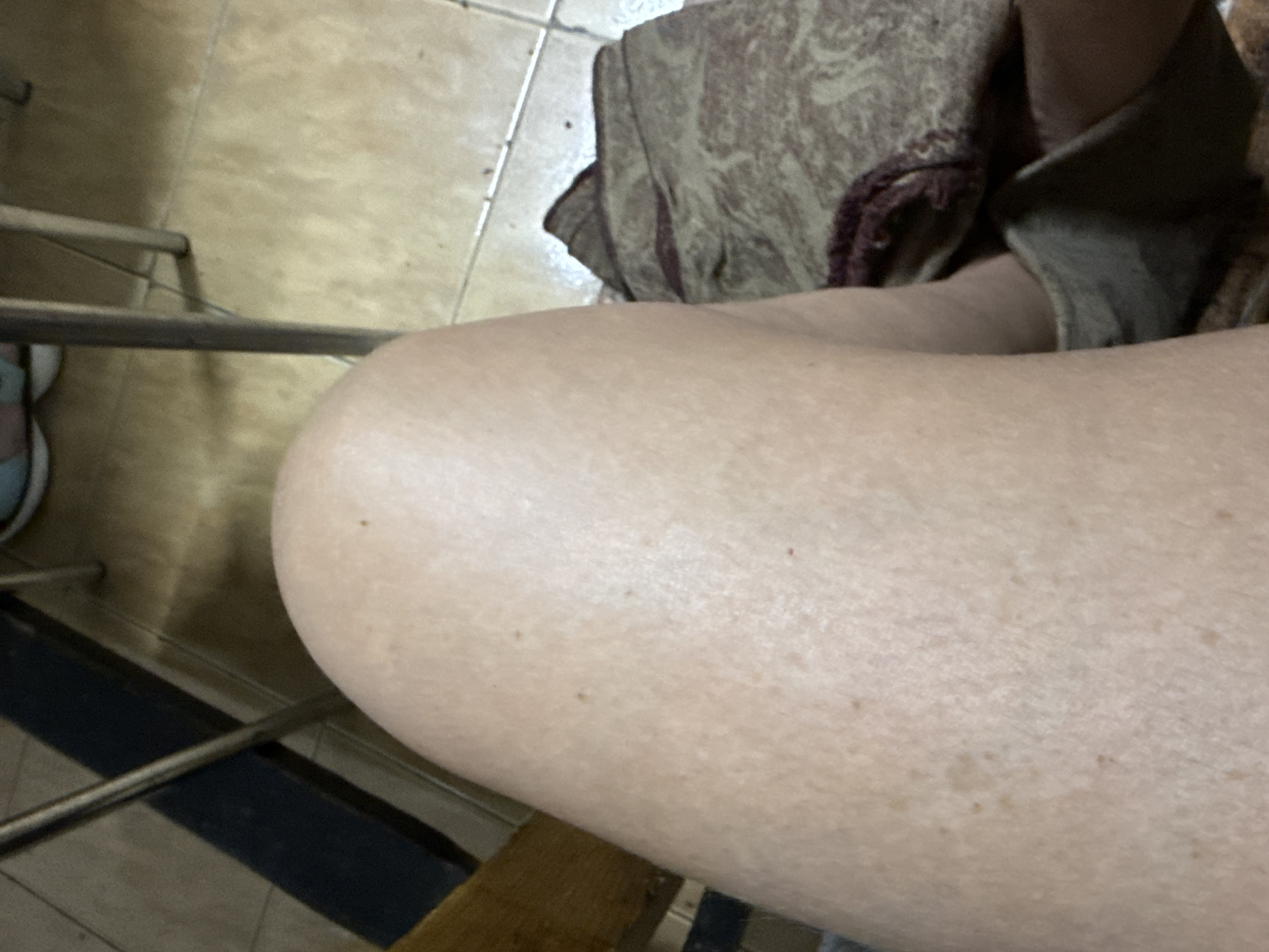 The image shows a person's leg in the foreground, with the person sitting on a chair with a metal frame. A folded cloth or garment is visible in the upper right of the frame, and the floor is tiled. There is no information present to indicate the location is Syniak, Ukraine, or any city.