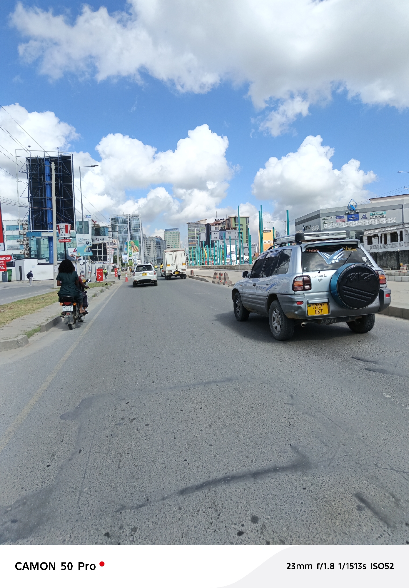 The image depicts a street scene in Dar es Salaam, Tanzania, on a sunny day with scattered clouds. In the foreground, there is a road with visible wear and tear. A silver Toyota RAV4 is positioned on the right side of the road, with its license plate "T782 DKT" clearly visible. Further down the road, a white sedan and a white truck are seen driving. On the left side of the road, a person is riding a motorcycle. In the background, there are buildings, including one with a "KFC" sign and another with a "TILE & STYLE" sign. There are also billboards and traffic barriers. The sky is blue with white clouds. The camera used to take the image is a "CAMON 50 Pro," and the image was taken with settings of 23mm f/1.8 1/1513s ISO52.