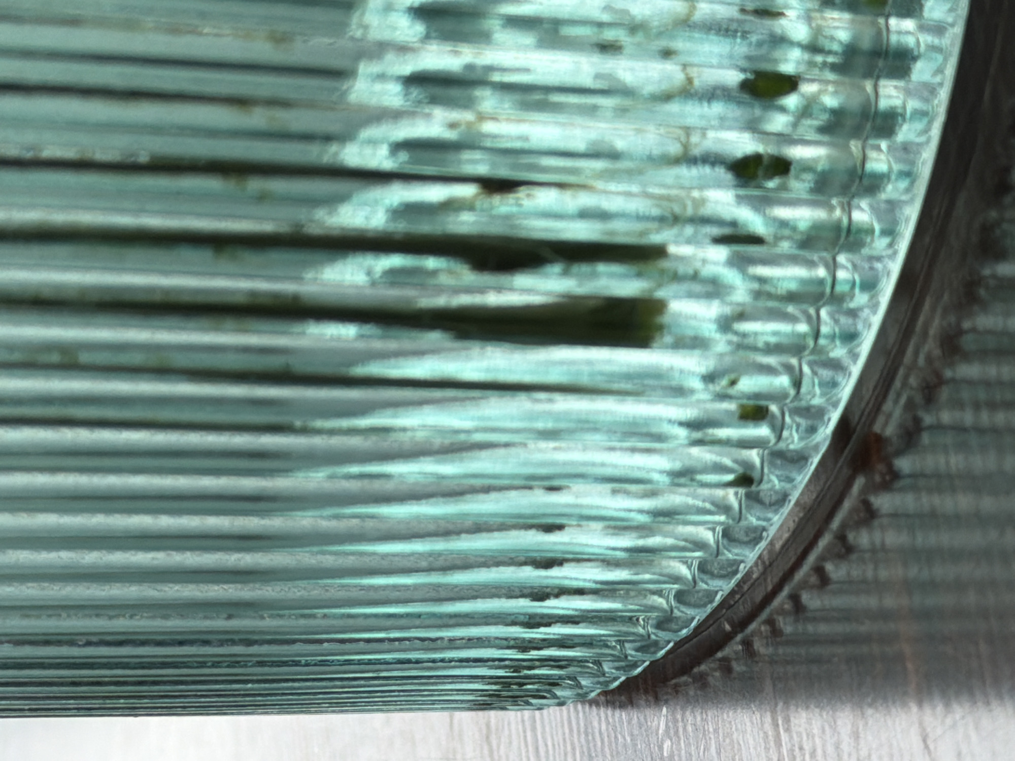 A close-up shot depicts a cylindrical, ridged, translucent teal glass element, possibly from the Moscow Metro. Dark residue or discoloration is visible within the glass ridges. The glass is set in a dark metal frame. A metallic surface reflects light in the lower right quadrant of the image. The shot focuses on the intersection of the glass, frame, and reflective surface.