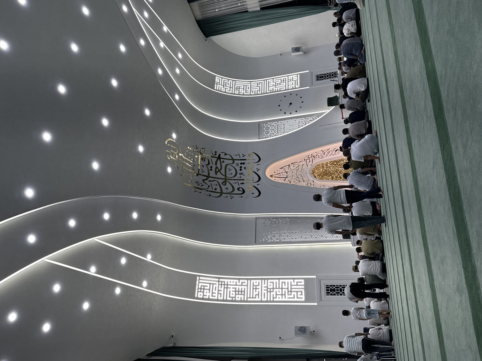 The media file shows a group of men praying in a mosque. They are all facing the same direction, likely towards the Kaaba in Mecca, as is customary in Islamic prayer. The mosque has a modern design with white, curved walls and intricate lighting. Golden Arabic calligraphy adorns the walls, adding to the spiritual ambiance. A clock on the wall indicates the time. The floor is covered with a green carpet, typical for mosques. The scene suggests a midday prayer, given the lighting conditions. The men appear focused and reverent. The location context of Tashkent, Uzbekistan, further grounds the image in a specific cultural and religious setting. The image captures a moment of communal worship, highlighting the devotion and unity of the Muslim community.