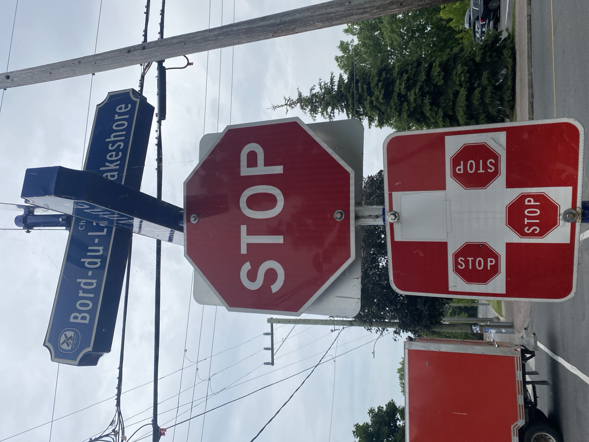 The image shows a street signpost at the intersection of Bord-du-Lac and Lakeshore in Pointe-Claire, Canada. The signpost has two blue directional signs pointing in opposite directions, indicating "Bord-du-Lac" and "Lakeshore." Below the directional signs, there is a large red octagonal stop sign with the word "STOP" in white letters. Further down the post, a smaller red and white sign displays three smaller octagonal stop signs. To the left, a red trailer truck is partially visible. The background features trees, utility poles, and power lines, with a cloudy sky overhead.