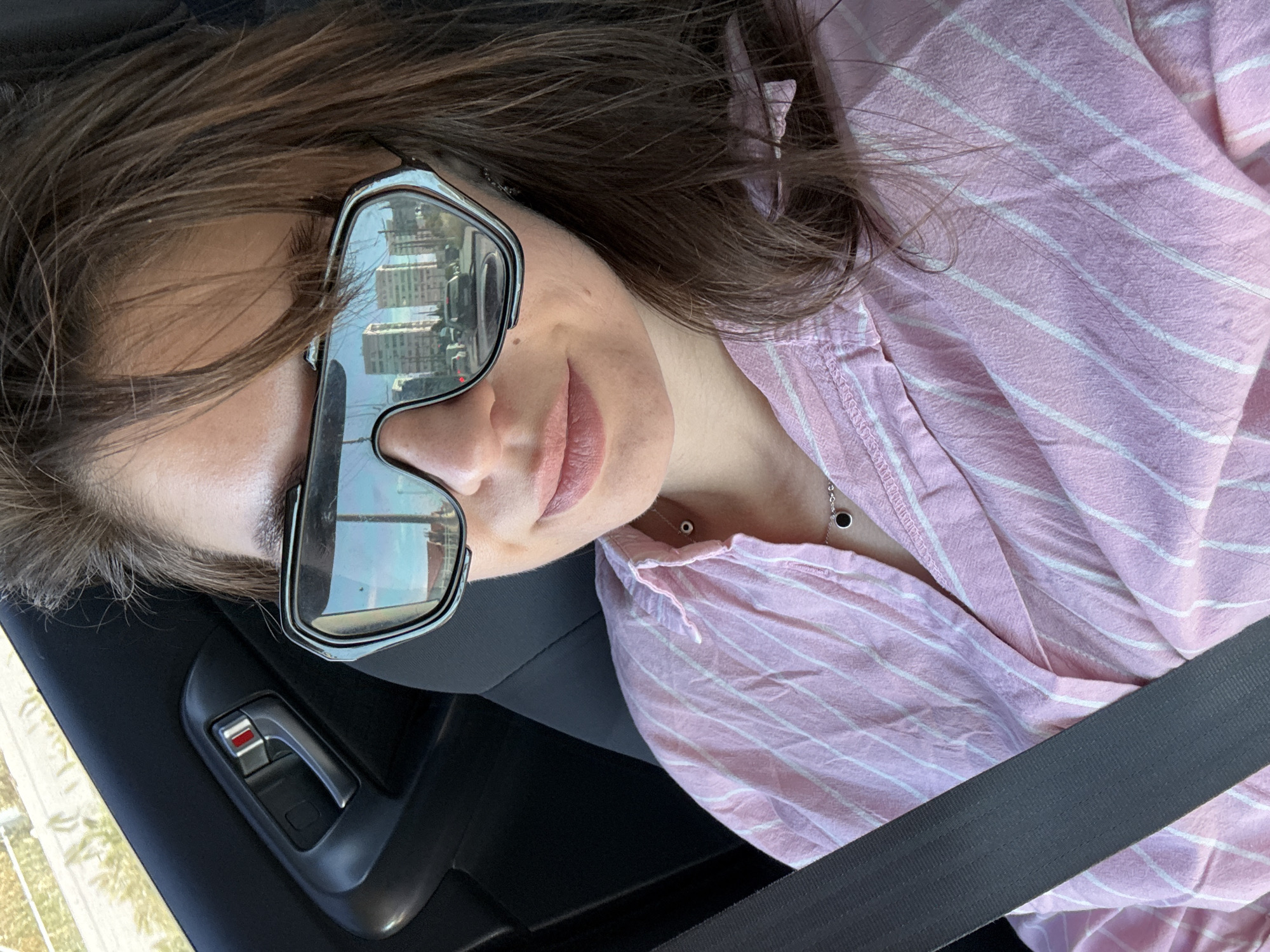 This is a close-up, angled, selfie-style photograph of a person wearing sunglasses and a pink and white striped shirt. The person is in the driver's seat of a car, evidenced by the car door handle and the seatbelt prominently displayed. The sunglasses are large and reflective, showing a distorted reflection of buildings and trees, suggesting an urban or suburban environment. The person's hair is dark brown and falls around their face and shoulders. They are wearing subtle makeup, with a touch of lip color. A delicate necklace with a small black pendant is visible at their collarbone. The lighting suggests it is daytime, possibly late afternoon, given the warm tones and the way the light falls on the clothing. The image is oriented such that the person appears to be looking out of the frame to the right. There is no visible text in the image.