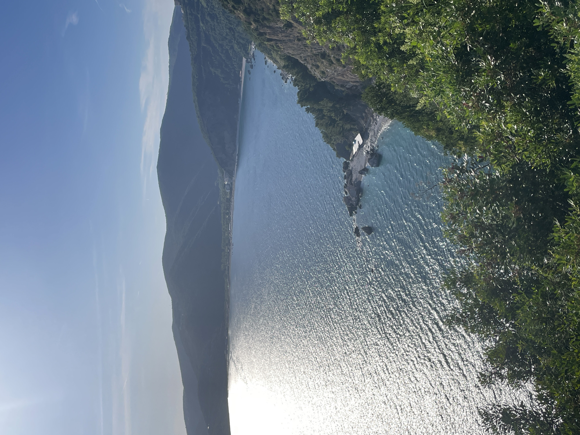The image shows a coastal scene in Budva, Montenegro, featuring a mountainous shoreline, a blue body of water reflecting sunlight, and lush green vegetation. The water appears to be the Adriatic Sea. A rocky outcropping extends into the water, with a small structure near the shore. The sky is clear and blue.