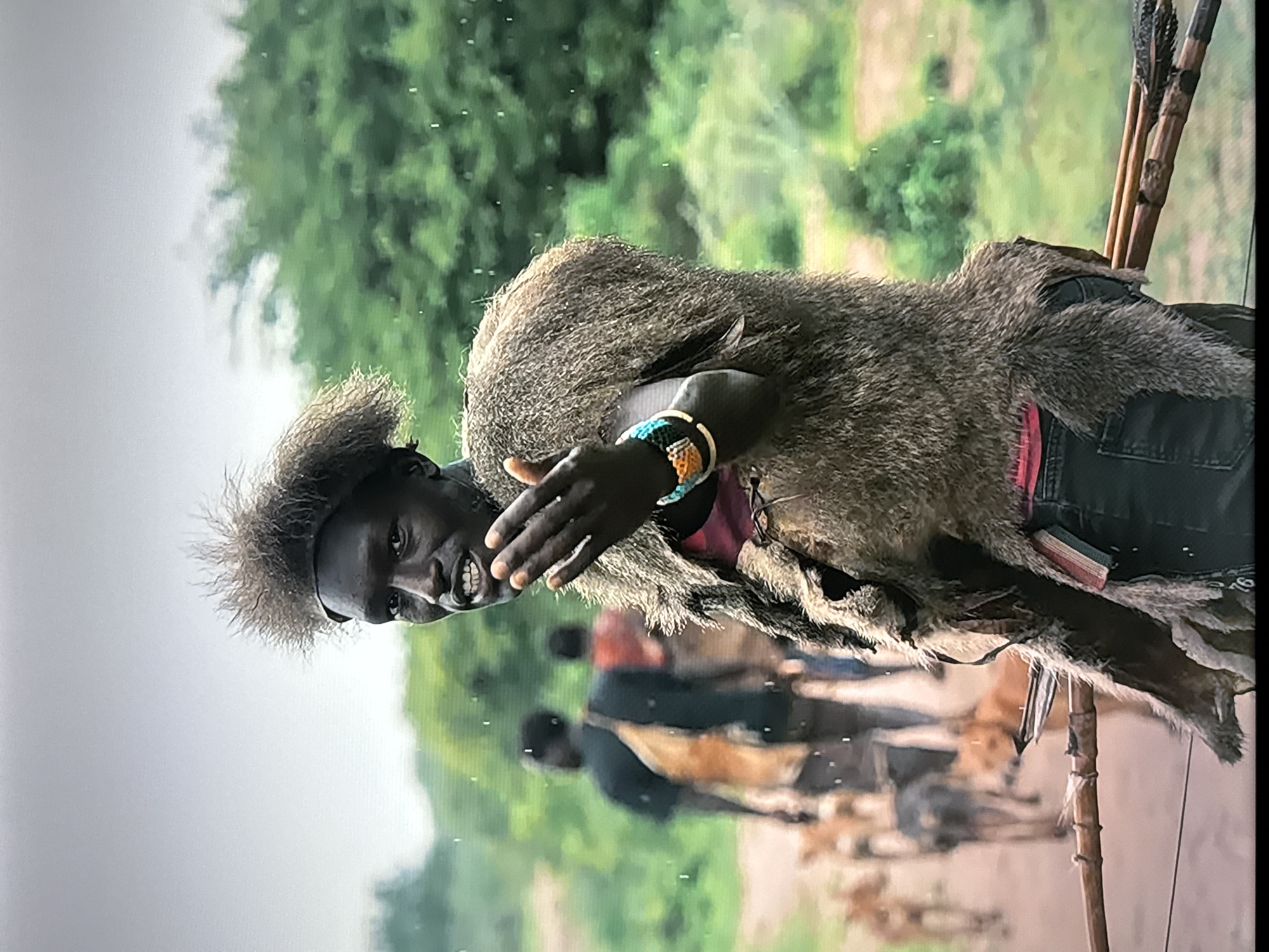A medium shot shows a man with dark skin and a spiky hairstyle standing in what appears to be a rural, possibly African, environment. He is wearing a brown, fur-like garment over a red undershirt and dark pants. The man is holding his left hand up and extended towards the camera, with his palm facing outwards. He is also wearing a colorful beaded bracelet on his left wrist. In the background, other people and dogs are visible, walking along a dirt path. Lush green trees and foliage are present in the background, and the sky appears overcast. The man is also holding a bow and arrows.